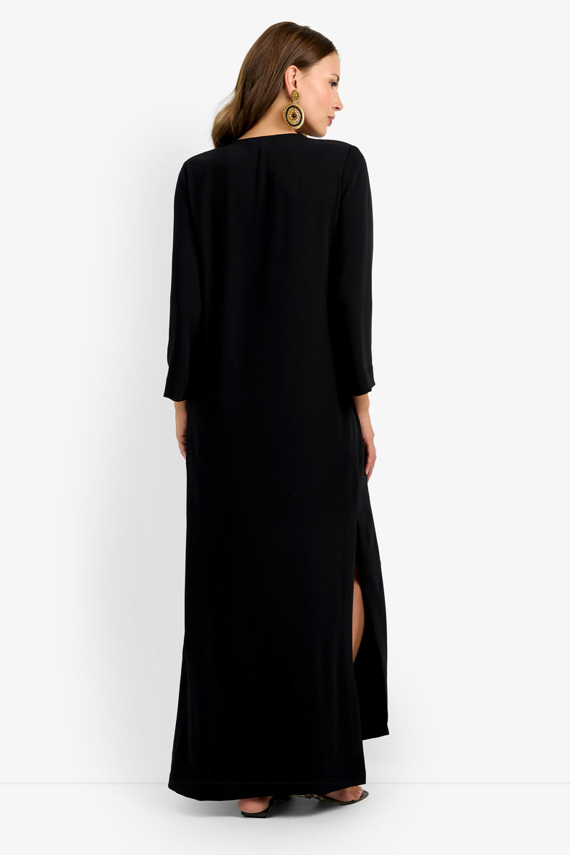 A woman with wavy brown hair and large circular earrings stands facing away, showcasing the Catamaran Dress—a long black, long-sleeved dress featuring a high side slit.
