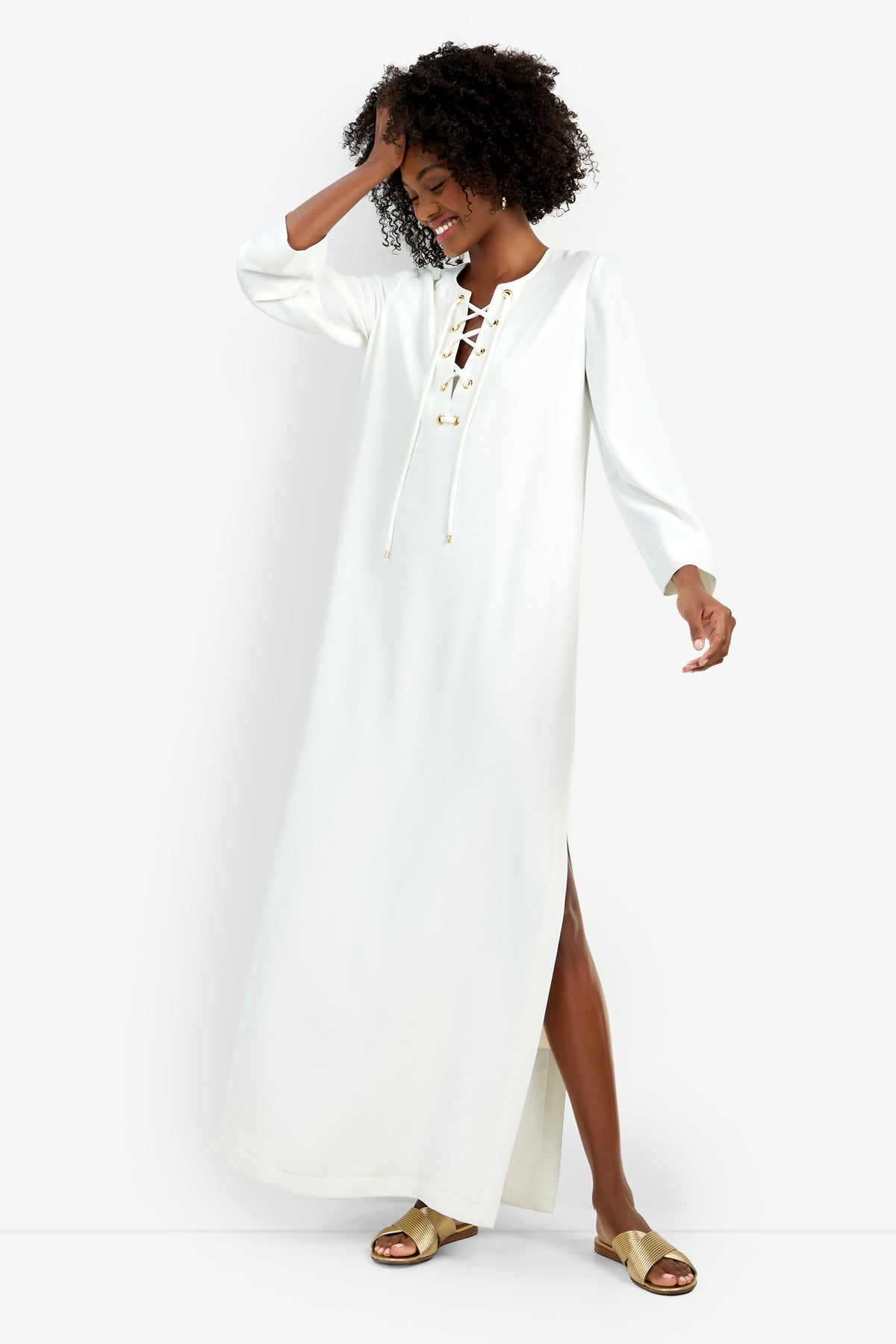 A woman smiles while wearing the Catamaran Dress, a long white dress with a lace-up neckline and long sleeves. She touches her hair with one hand, styled with gold sandals, and poses against a plain white background.