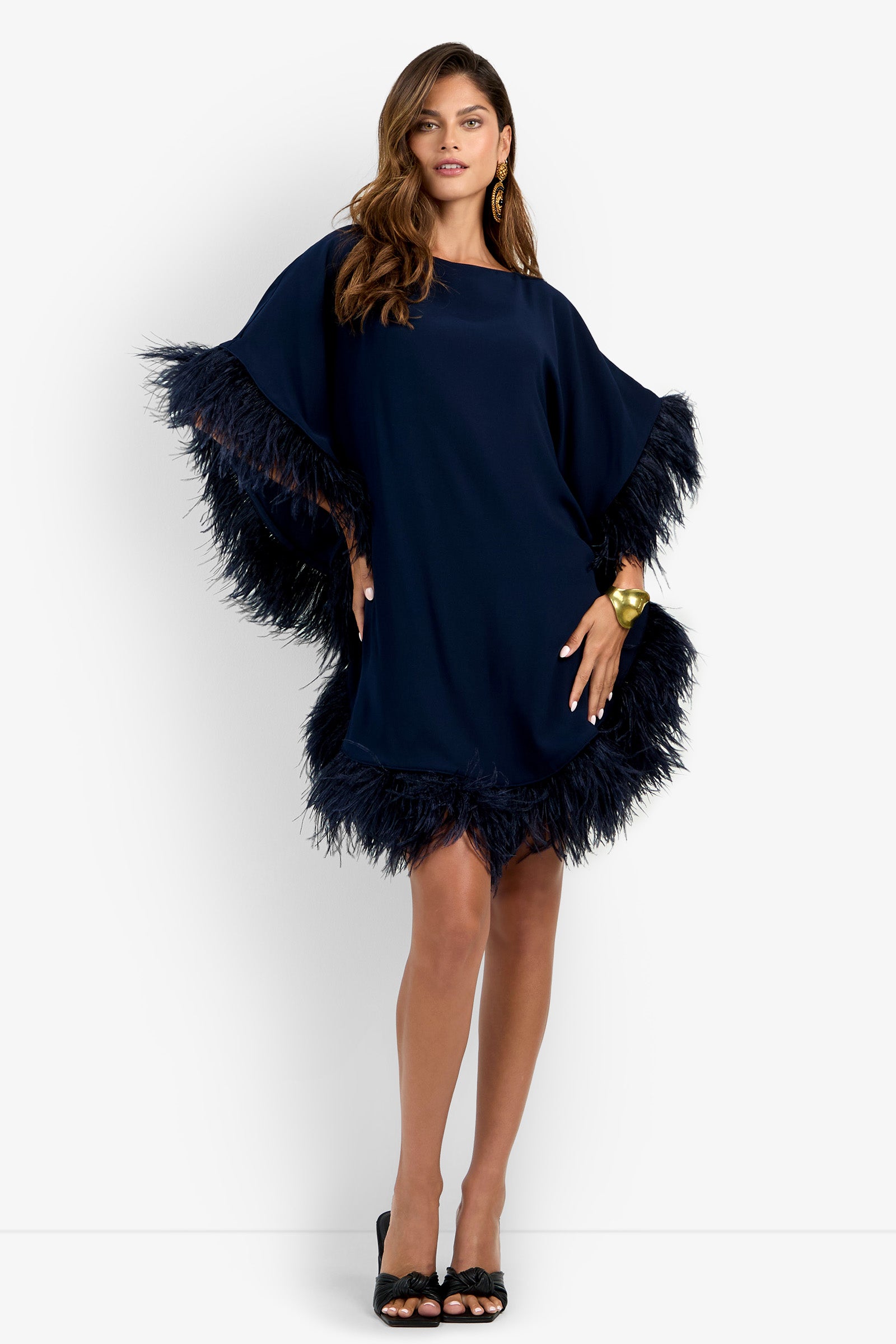 A woman stands with one hand on her hip and the other relaxed, wearing the Mini Neena Fringe Dress in navy blue, featuring feathered trim on the sleeves and hem. She completes the look with black heels and a gold bracelet.