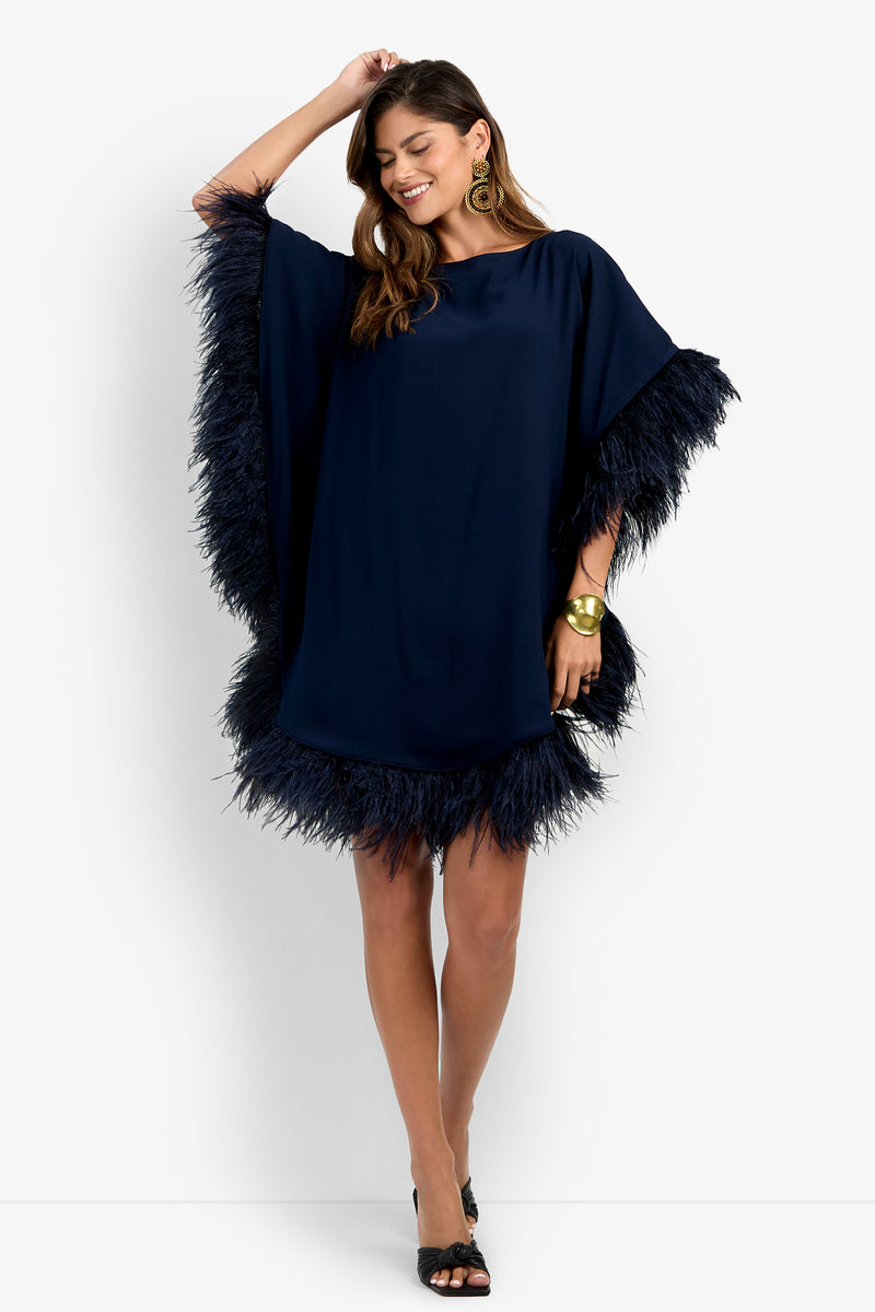 Smiling woman wearing the Mini Neena Fringe Dress in navy blue with feathered trim on the sleeves and hem, paired with black open-toe heels and a large gold bracelet, posing with one hand in her hair.