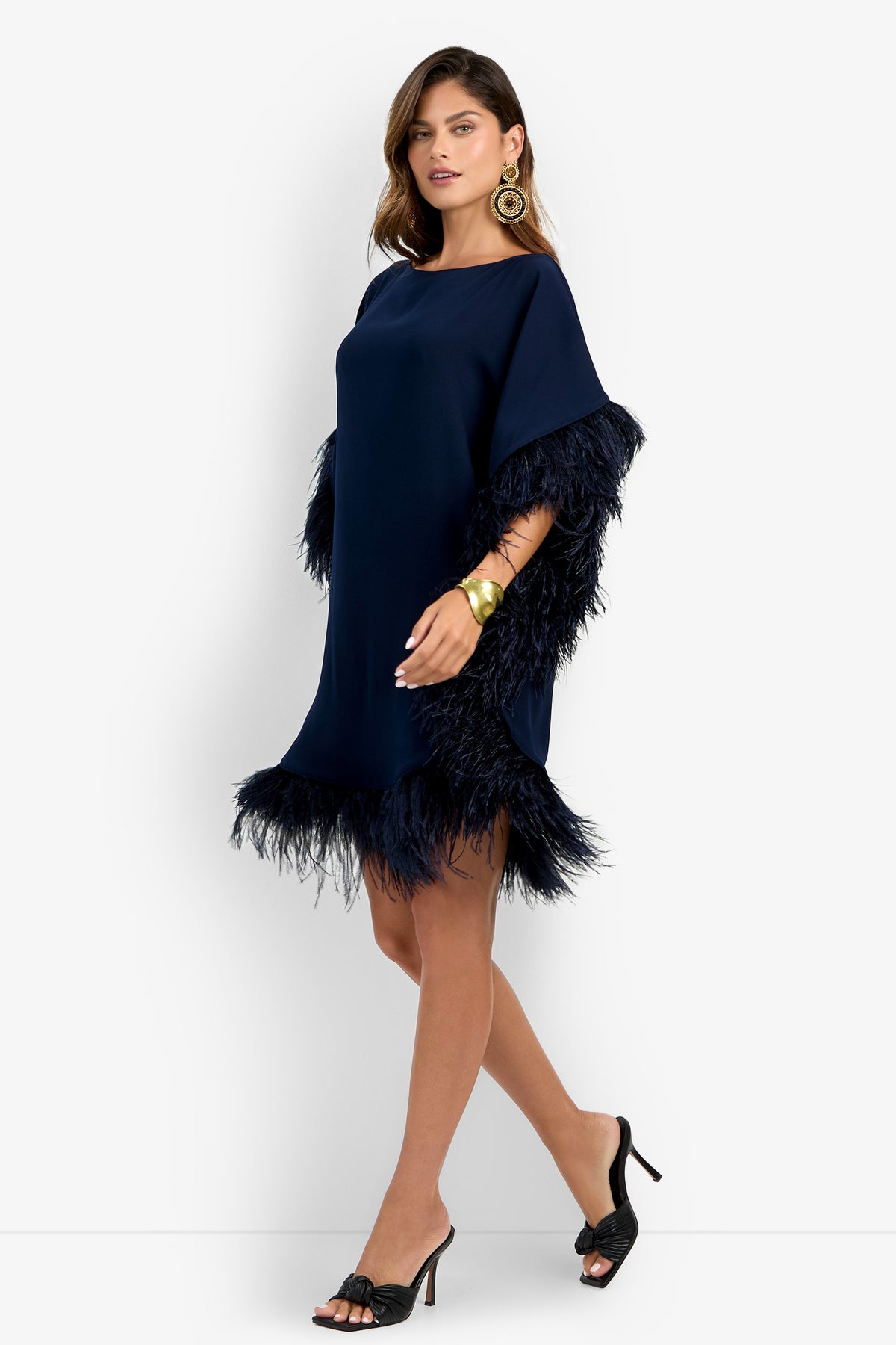A woman wearing the Mini Neena Fringe Dress, featuring navy blue fabric with feather trim on the sleeves and hem, paired with black heeled sandals, large gold earrings, and a gold bracelet, poses against a plain white background.