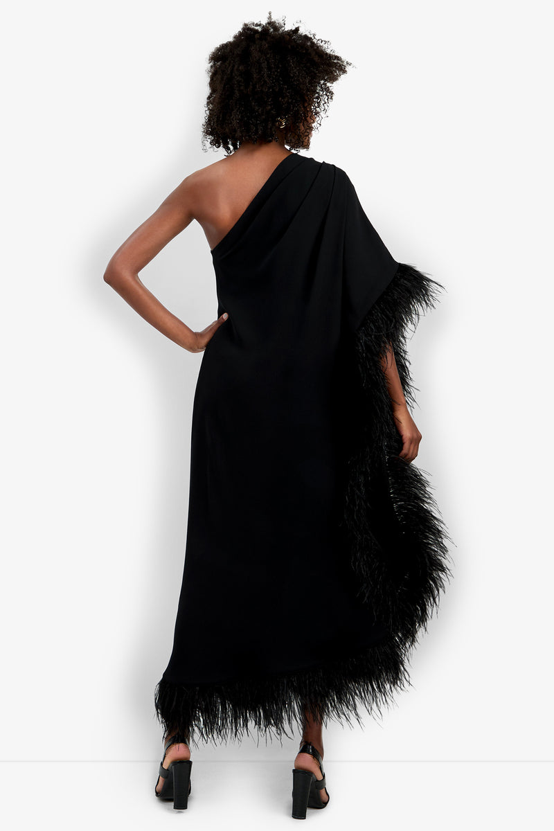 A person with curly hair stands facing away in a black Largo Caftan featuring a one-shoulder design and feathered trim along the hem and edge, paired with black strappy heeled sandals, against a plain white background.