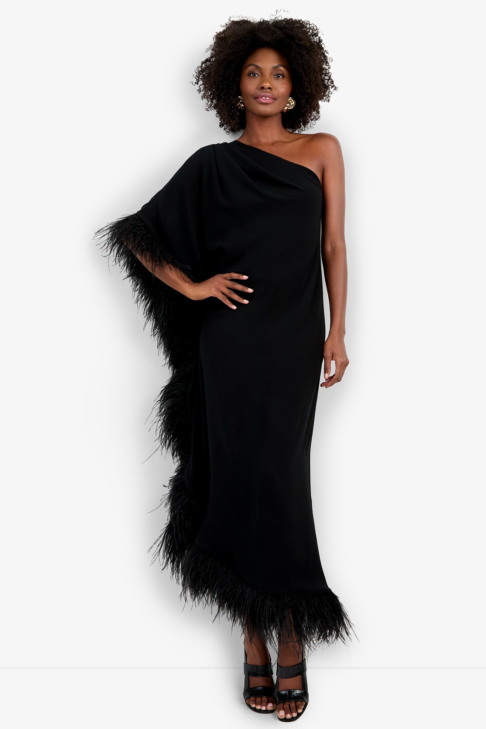 A woman with curly hair poses against a white background in the Largo Caftan, a one-shoulder black gown featuring feather trim at the edge and hem. She completes her look with black heeled sandals and stands with one hand on her hip.
