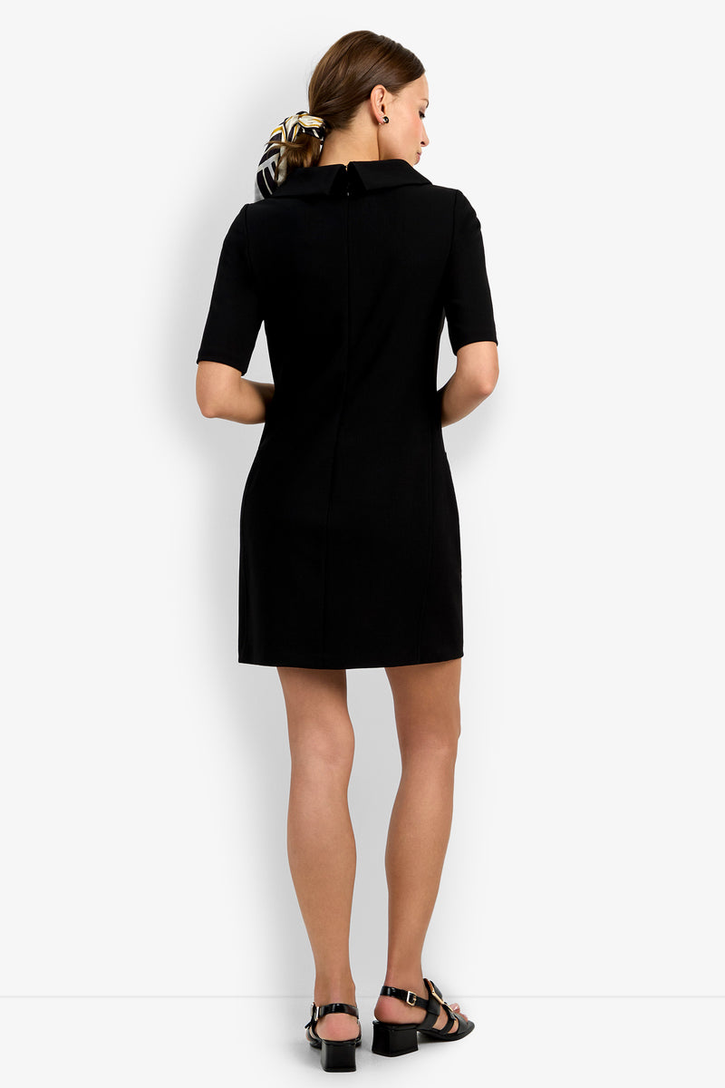 A woman faces away in the Maleko Dress, a short-sleeve black dress, paired with black sandals and a black-and-white patterned scrunchie. Her hair is tied back against a plain white background.