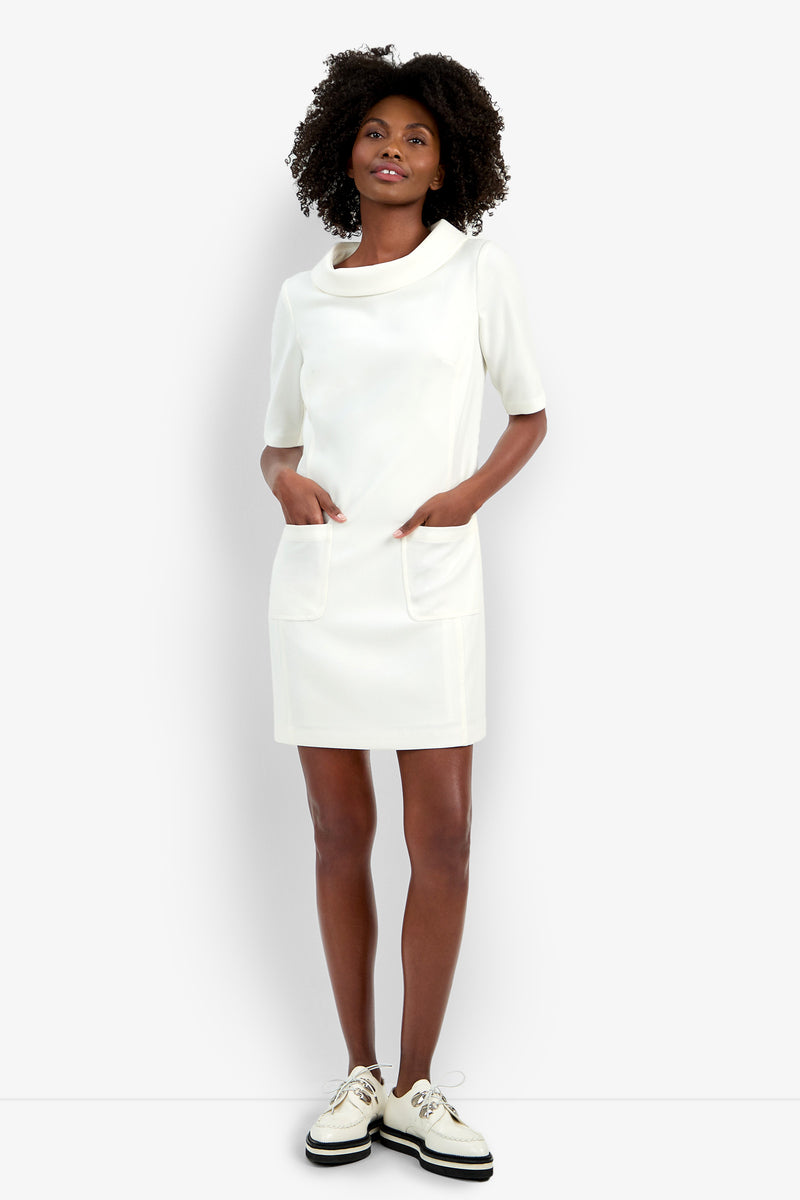 A woman with curly hair smiles slightly while wearing the Maleko Dress—a short-sleeved, knee-length white dress with front pockets—paired with white platform shoes, standing against a white background.