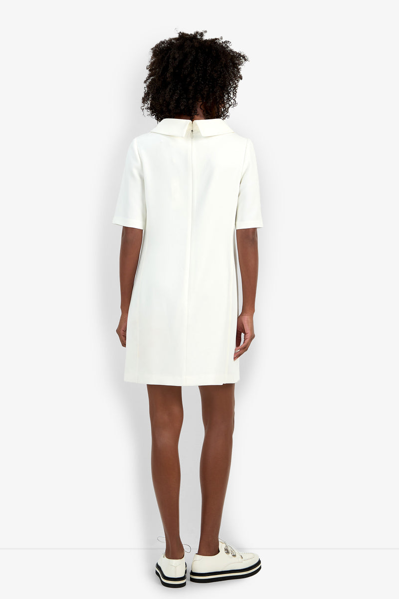 A person with curly hair faces away, wearing the Maleko Dress—a short-sleeved, collared white mini dress with a visible back zipper—paired with white platform shoes against a plain white background.