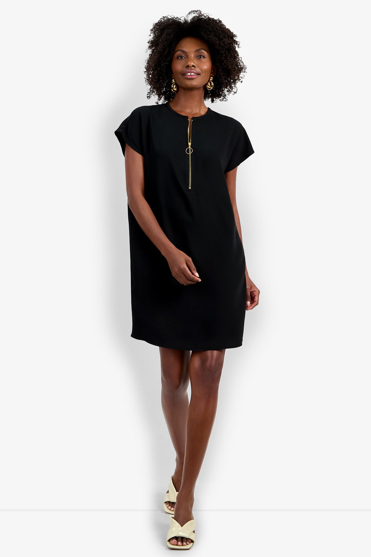 A woman smiles against a plain white background wearing the Hasil Dress, a short-sleeve black dress with a front zip detail, paired with beige open-toe heels.