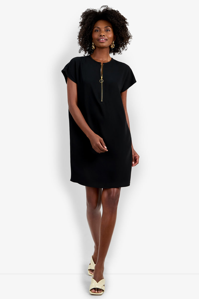 A woman smiles against a plain white background wearing the Hasil Dress, a short-sleeve black dress with a front zip detail, paired with beige open-toe heels.
