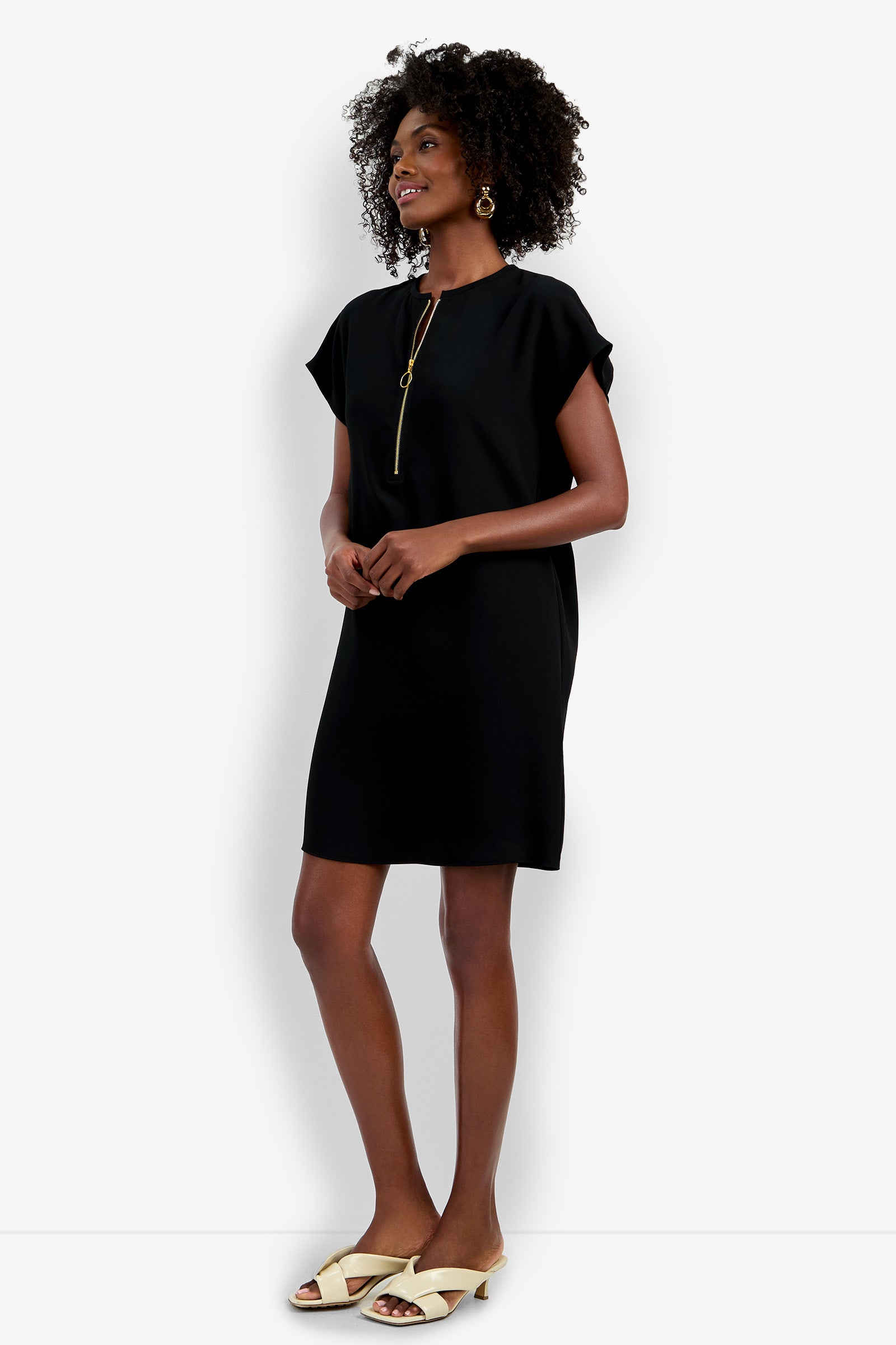 A woman with curly hair smiles in the Hasil Dress—a simple black, short-sleeve dress—styled with a gold necklace, hoop earrings, and white open-toe slides. She stands against a plain white background.