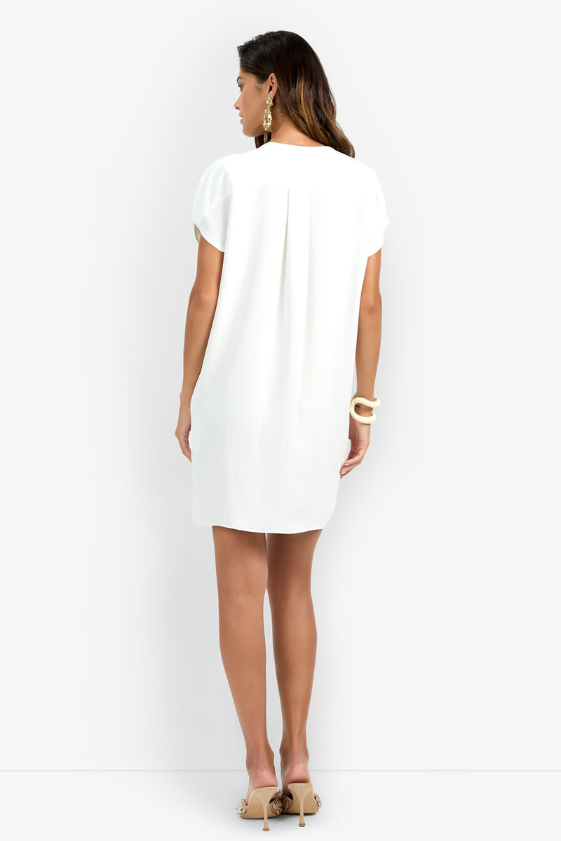 A woman stands with her back to the camera against a plain white background, wearing the Hasil Dress—a short-sleeved, knee-length white dress—paired with beige heels, statement earrings, and her long hair cascading down.