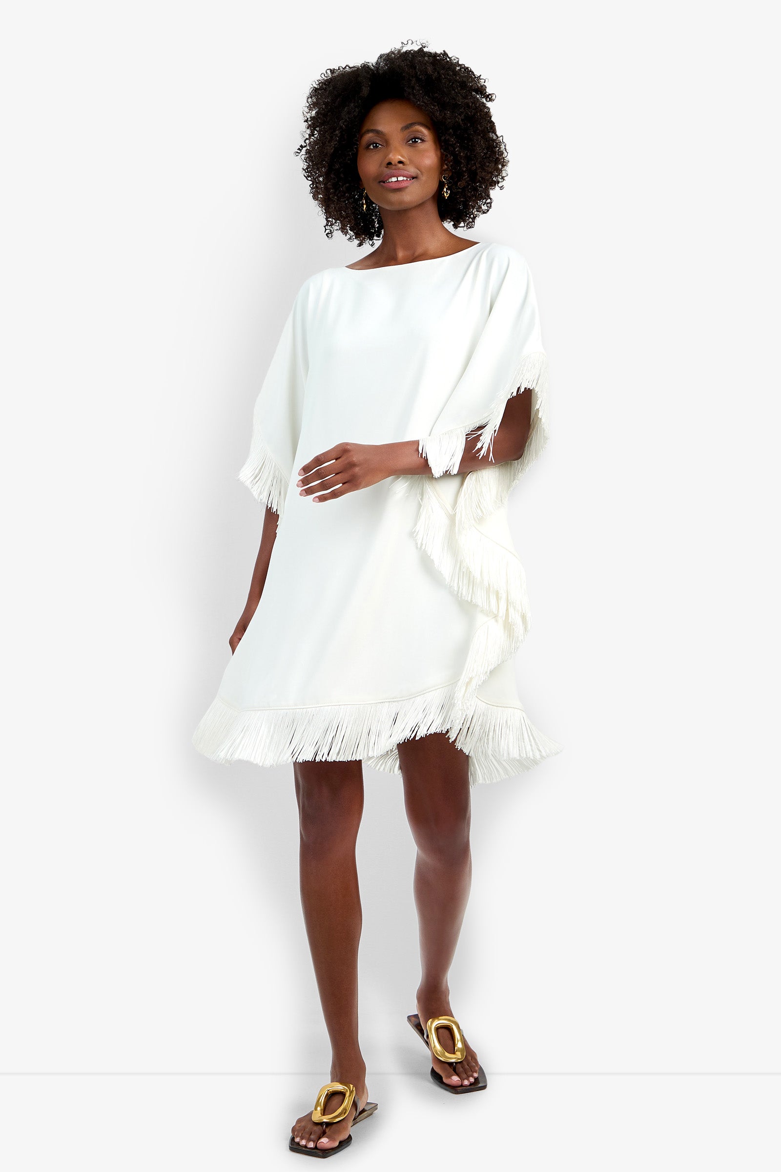 A woman wears the Mini Neena Fringe Dress, a loose-fitting white dress featuring fringe on the sleeves and hem, paired with flat brown sandals. She stands against a plain white background, smiling gently.