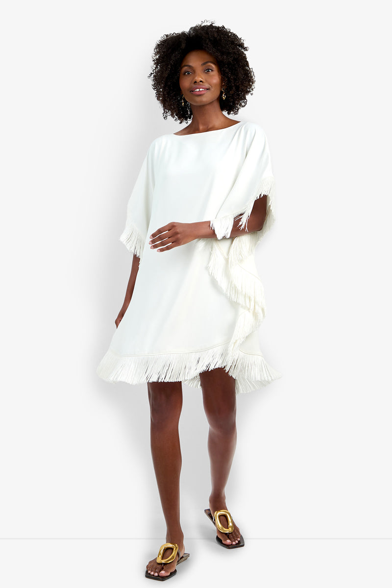 A woman wears the Mini Neena Fringe Dress, a loose-fitting white dress featuring fringe on the sleeves and hem, paired with flat brown sandals. She stands against a plain white background, smiling gently.