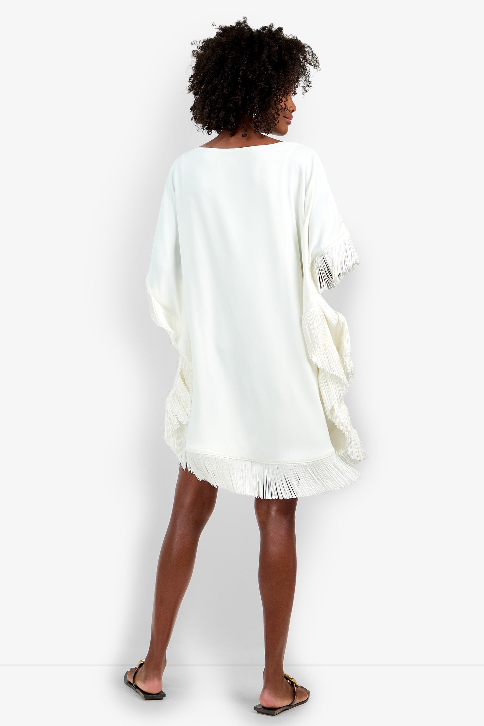 A woman with curly hair stands facing away against a white background, wearing the loose-fitting Mini Neena Fringe Dress featuring fringe details on the sleeves and hem, paired with flat sandals.