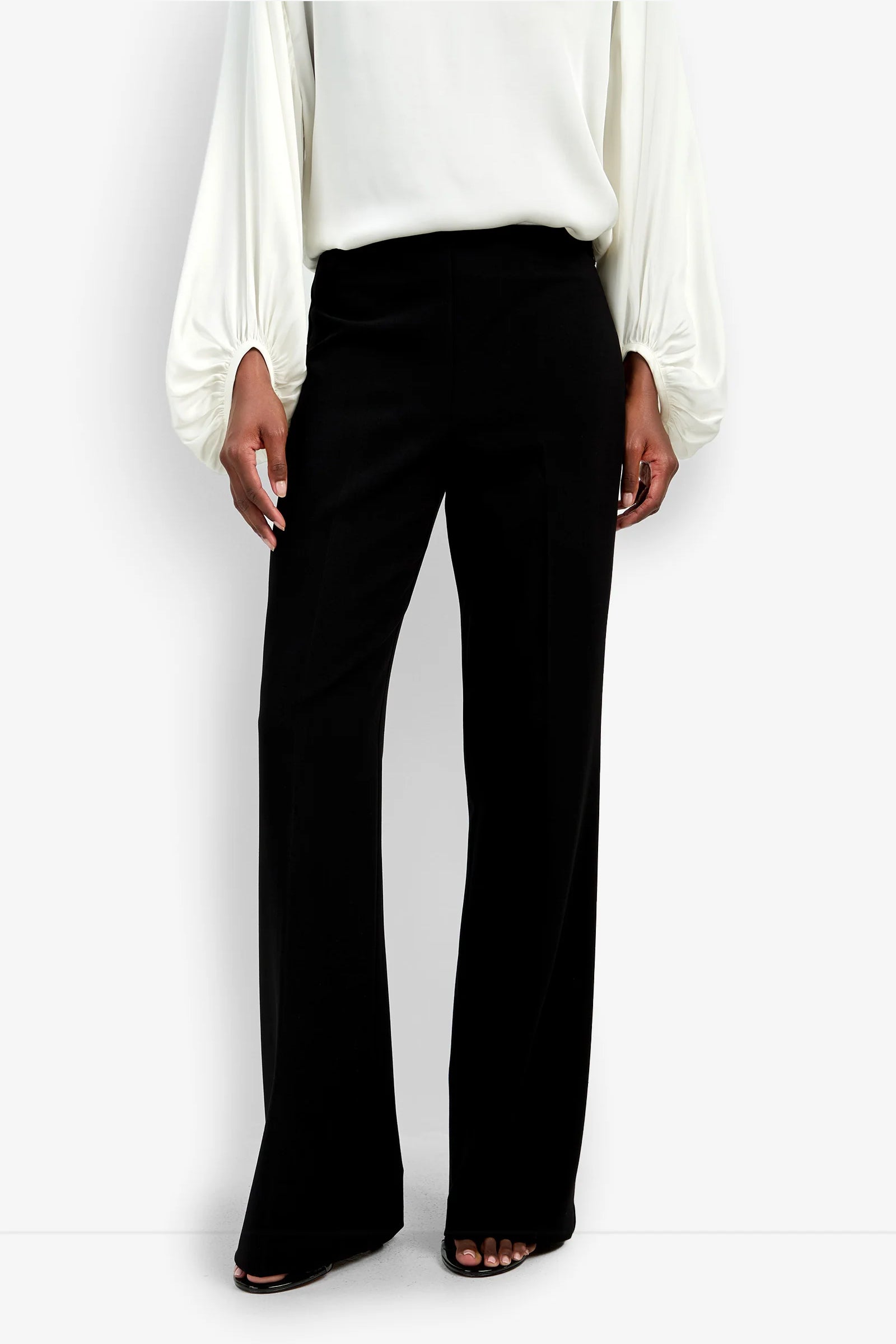 A person is wearing high-waisted, wide-leg black Chimayo Pants paired with a loose, long-sleeve white blouse. The image is cropped at the shoulders and feet to highlight the clothing style.