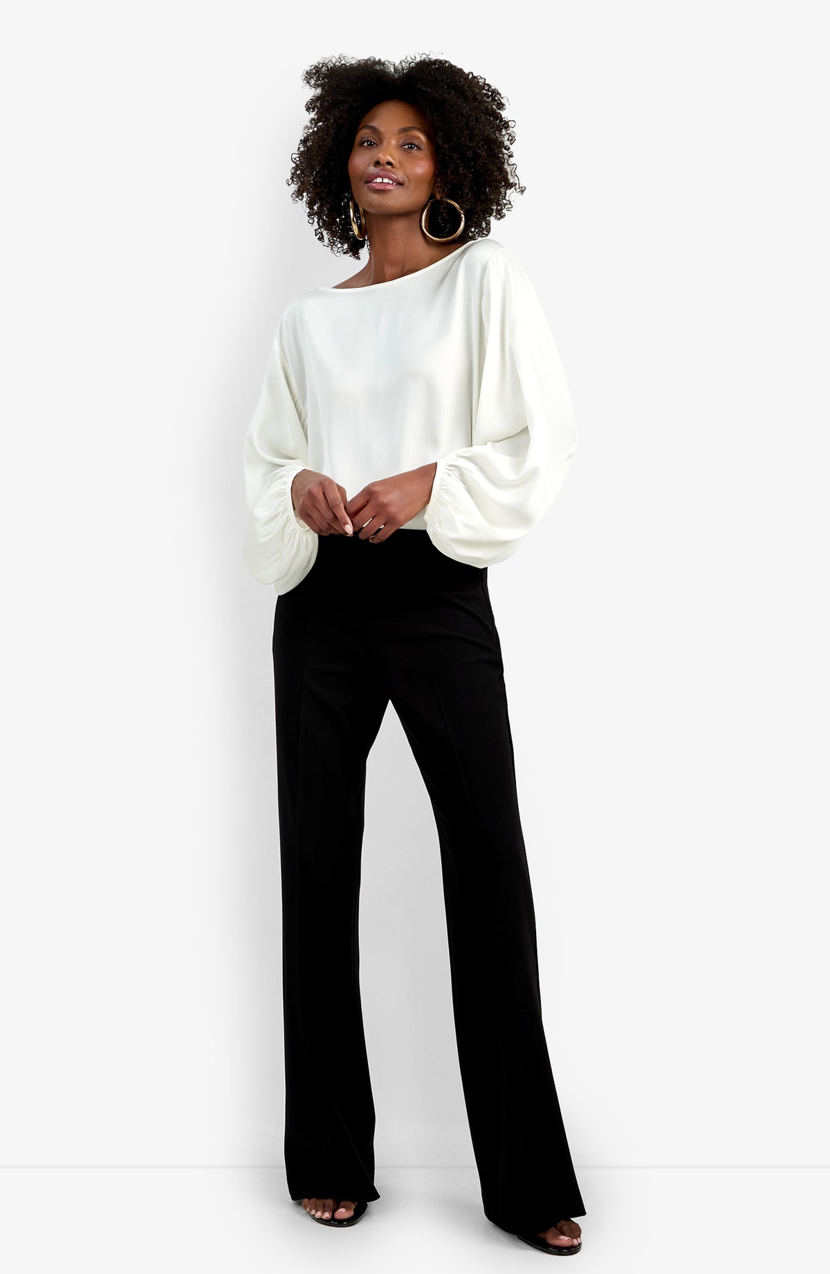 A woman smiles against a white background, wearing black Chimayo Pant wide-leg trousers, a loose white long-sleeve top with billowy sleeves, hoop earrings, and open-toed shoes. She stands with her hands lightly clasped.