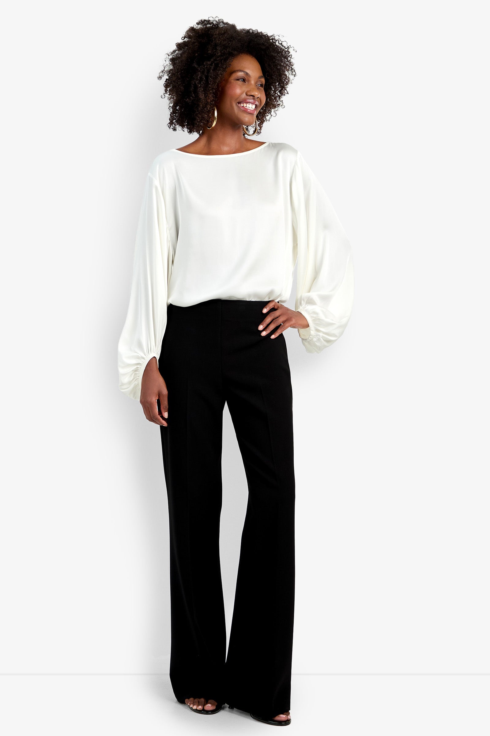 A woman smiles, wearing the Chimayo Pant—high-waisted, wide-leg black trousers—with a long-sleeved white blouse and black open-toe shoes. One hand rests on her hip.