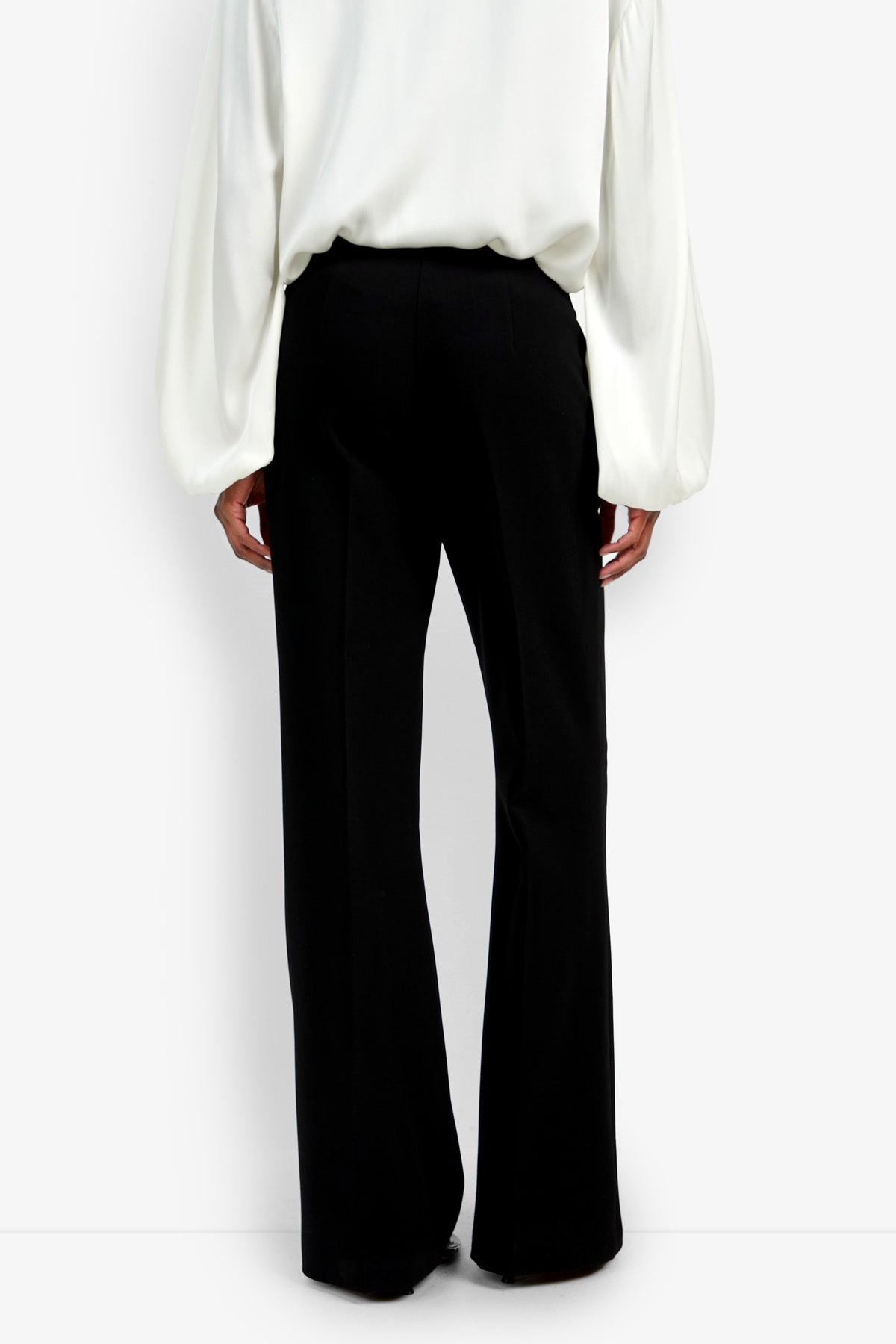 A person stands facing away, wearing the Chimayo Pant in black and a long-sleeved white blouse with billowy sleeves, set against a plain white background.