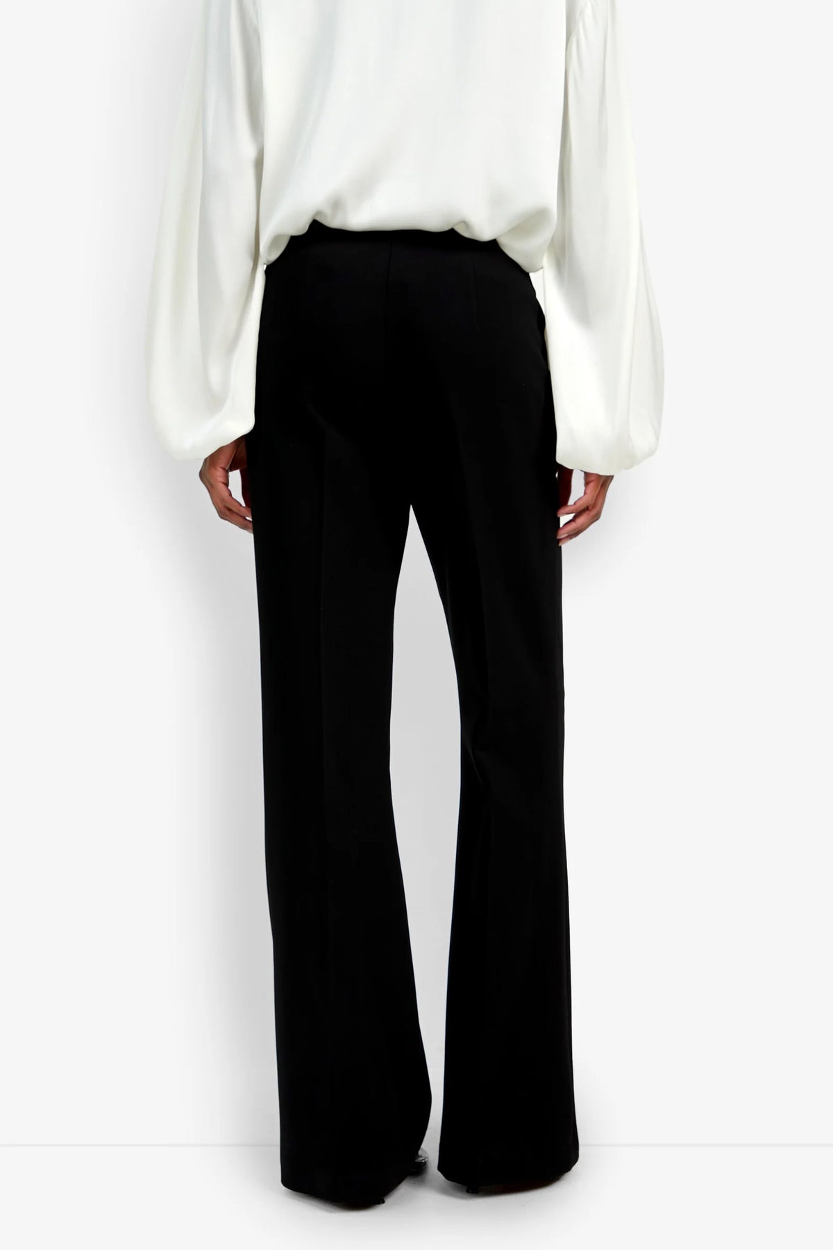 A person wearing the Chimayo Pant in black wide-leg style, paired with a long-sleeved white blouse, shown from the back against a plain white background.