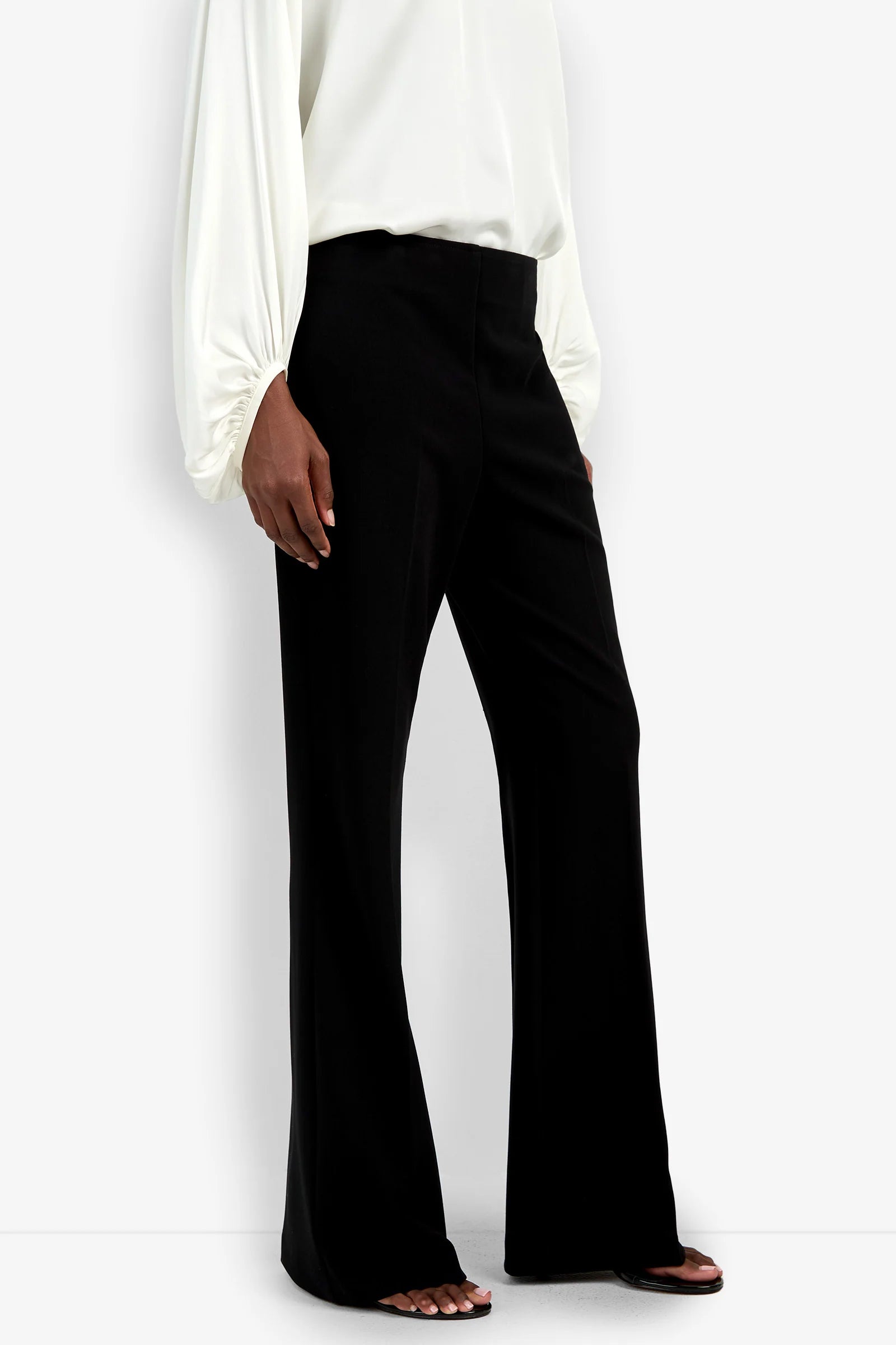 A person wearing the Chimayo Pant in black and a long-sleeved white blouse stands against a white background, with one hand relaxed by their side.