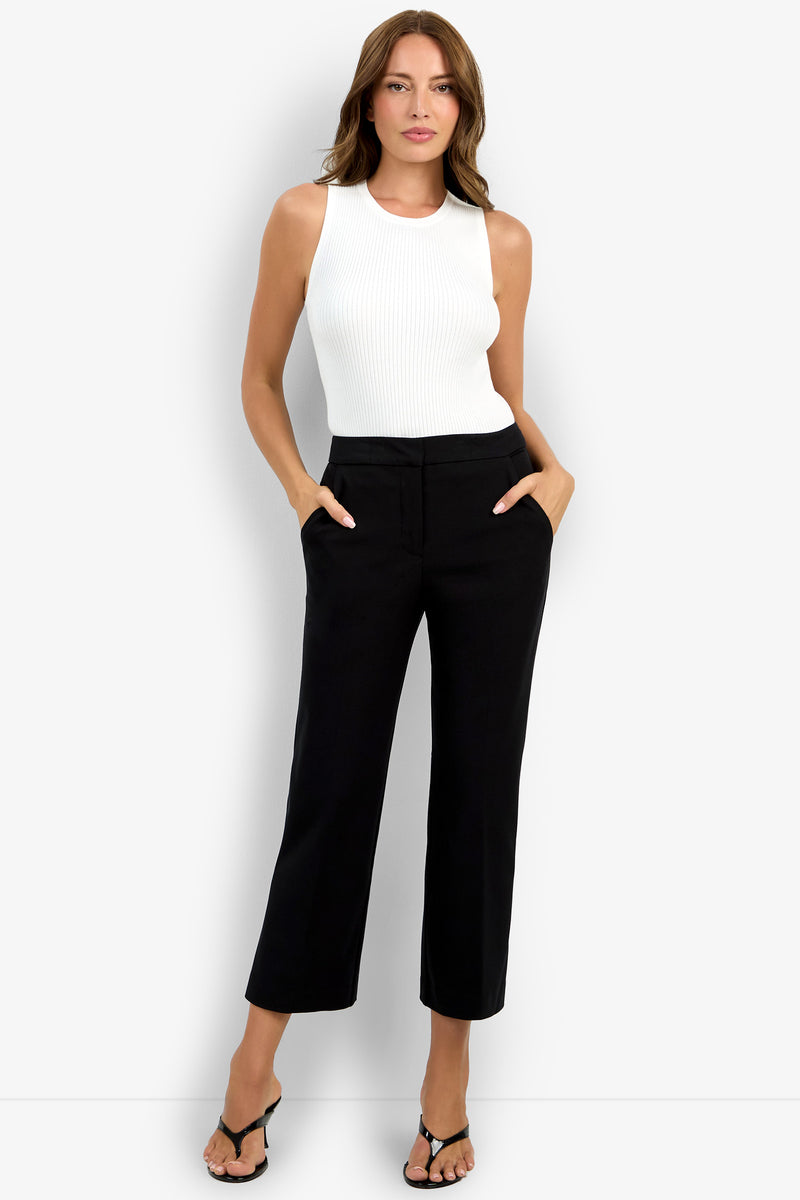A woman stands against a white background wearing black Lulu Pant high-waisted trousers, a sleeveless white ribbed top, and black heeled sandals. With her hands in her pockets, she gazes confidently at the camera.