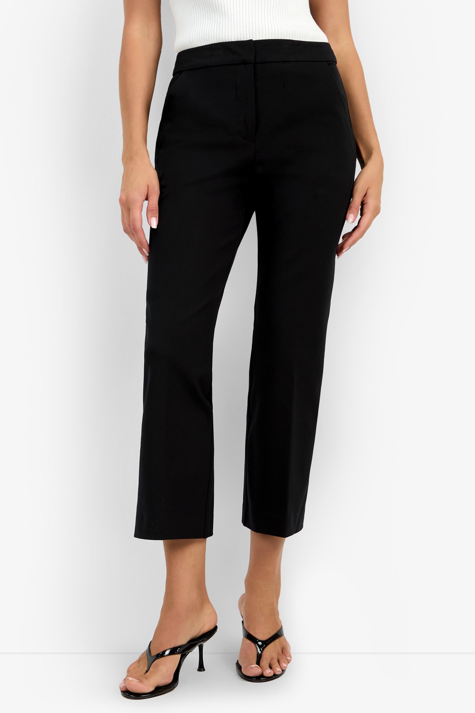 A woman models the Lulu Pant—black cropped dress pants—paired with a white sleeveless top and black heeled sandals, standing against a white background. Her arms are relaxed at her sides; her head is not visible in the photo.