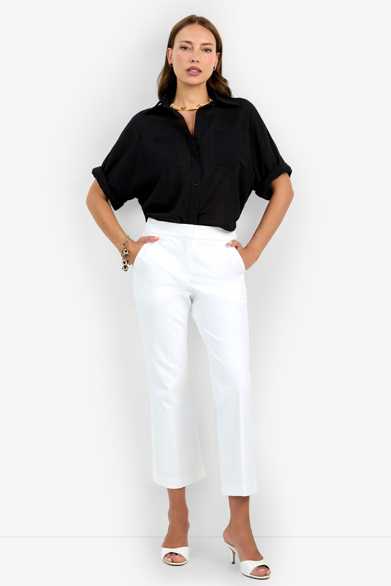 A woman stands against a white background, wearing the Lulu Pant in white with a black short-sleeve button-up shirt, white heeled sandals, and jewelry. Her hands are in her pockets as she looks at the camera.