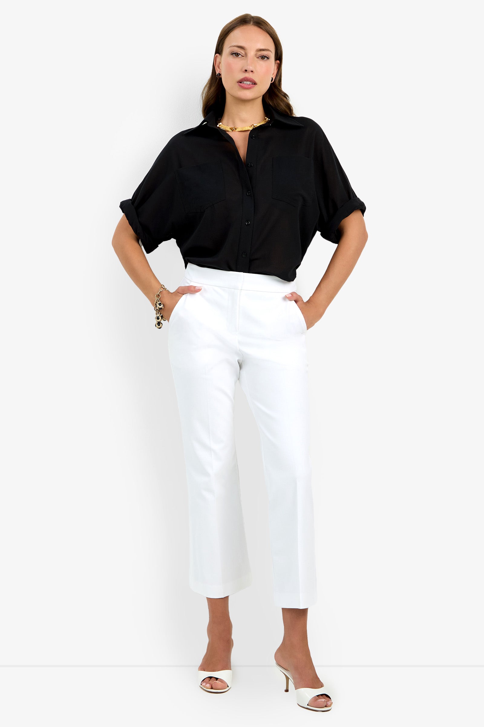 A woman stands with hands in her pockets, wearing a black short-sleeve button-up shirt tucked into high-waisted Lulu Pant in white and white heeled sandals against a plain background.
