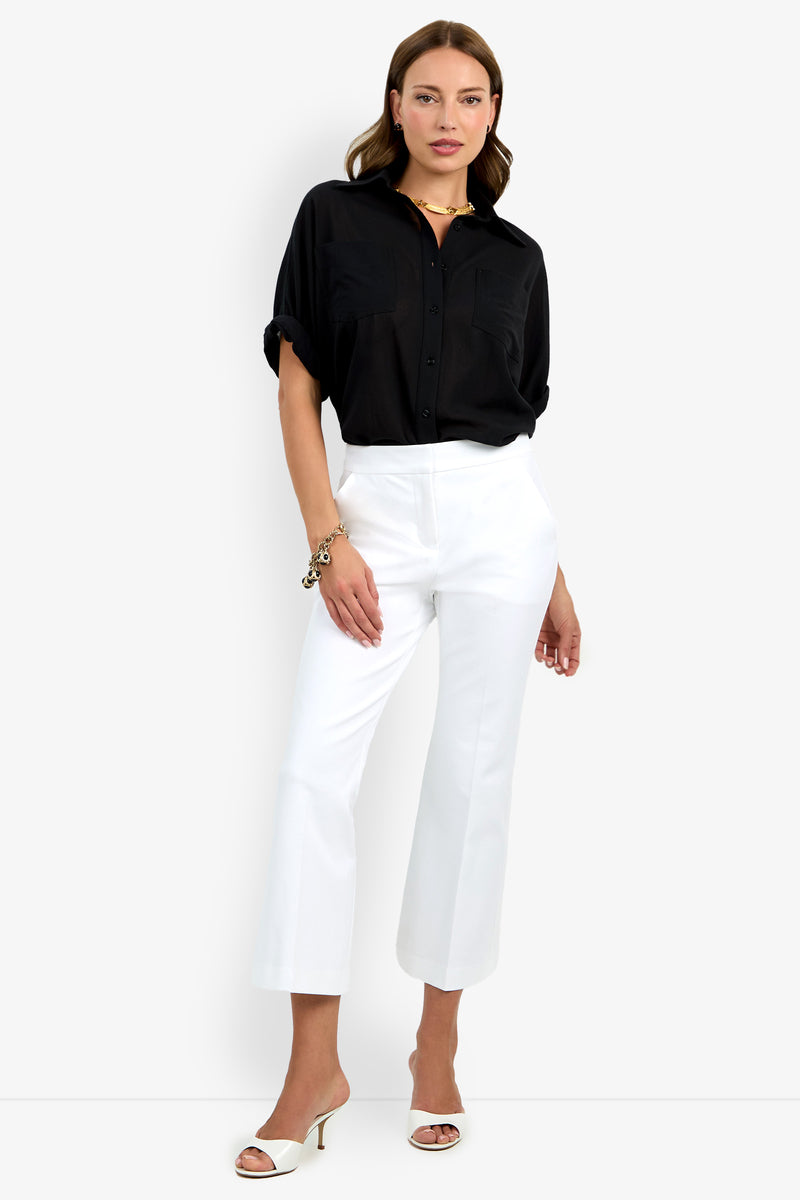 Woman standing against a plain background, wearing a black button-up shirt with rolled sleeves, Lulu Pant in white, white heeled sandals, and gold jewelry.