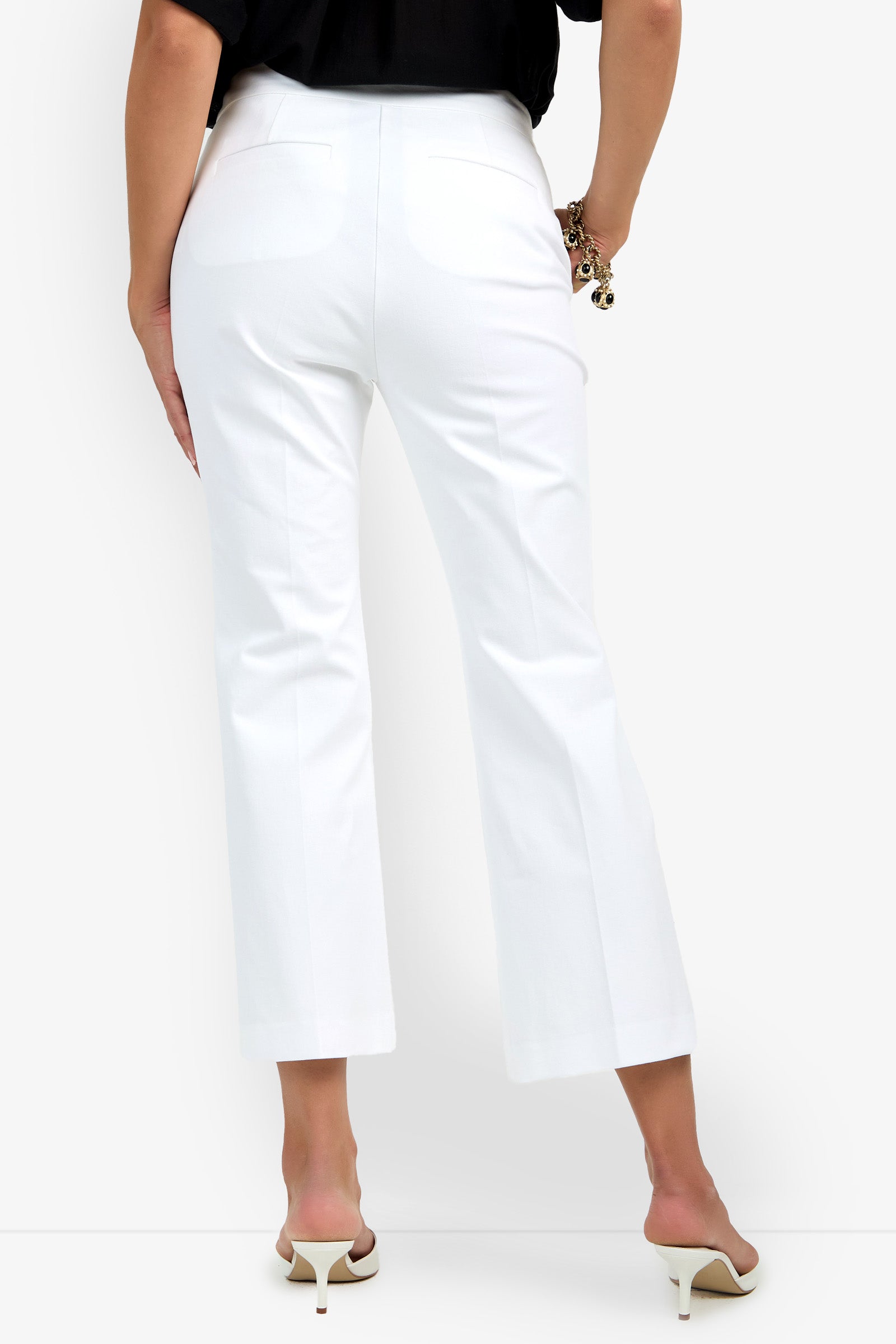 A person is seen from the back wearing Lulu Pant in white and white heeled sandals, with one hand on their hip and the other holding sunglasses, against a plain white background.