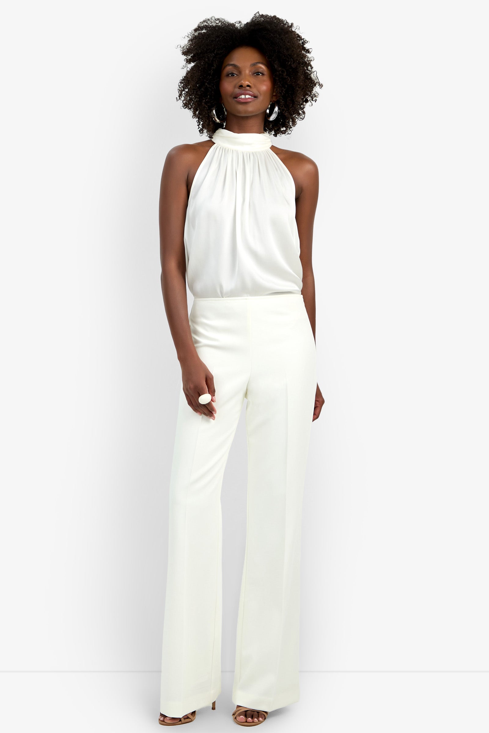 A woman with curly hair smiles confidently against a white background, wearing the Chimayo Pant—high-waisted, wide-leg white trousers—paired with a sleeveless high-neck blouse and open-toe heels.