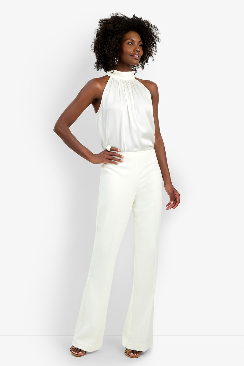A woman stands confidently, smiling with one hand on her hip, wearing high-waisted wide-leg white Chimayo Pants and a sleeveless white halter-neck blouse. She has curly hair and exudes effortless style.