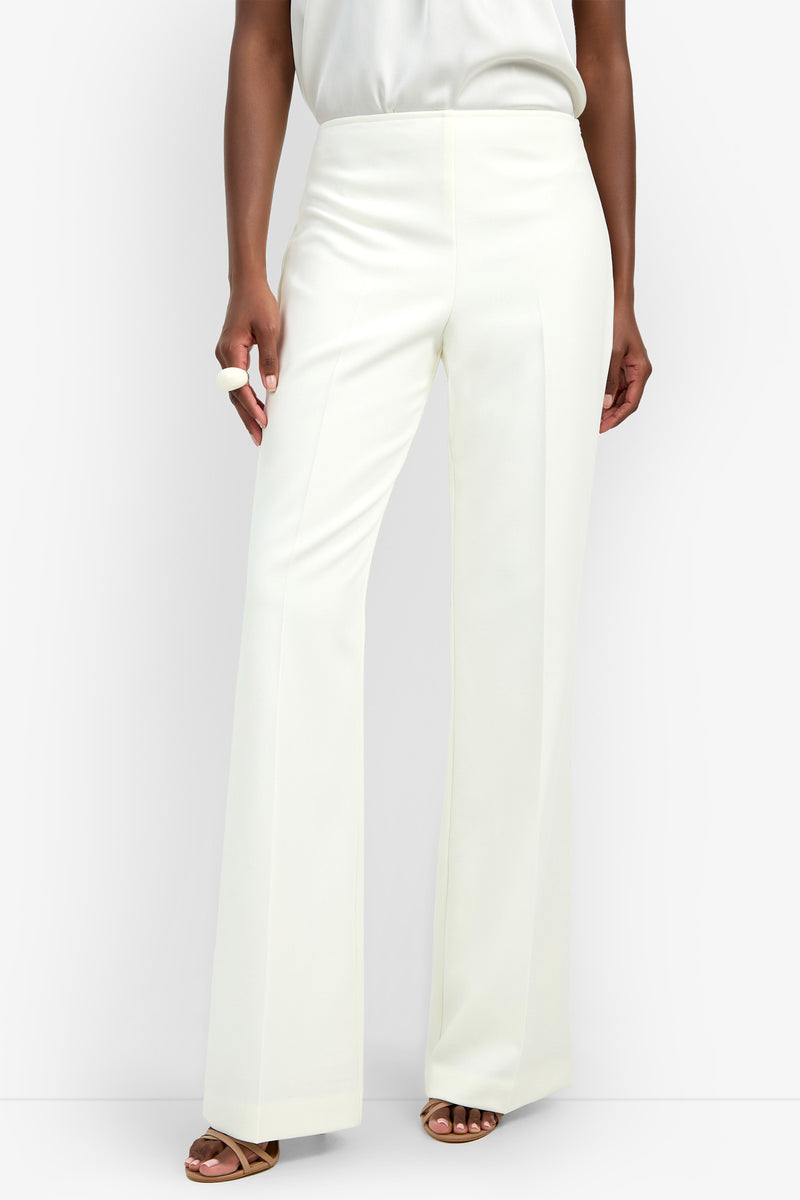 Someone stands against a plain background wearing the Chimayo Pant, which is high-waisted, wide-leg, and white, paired with a white top, brown heeled sandals, and a visible white ring on one hand.