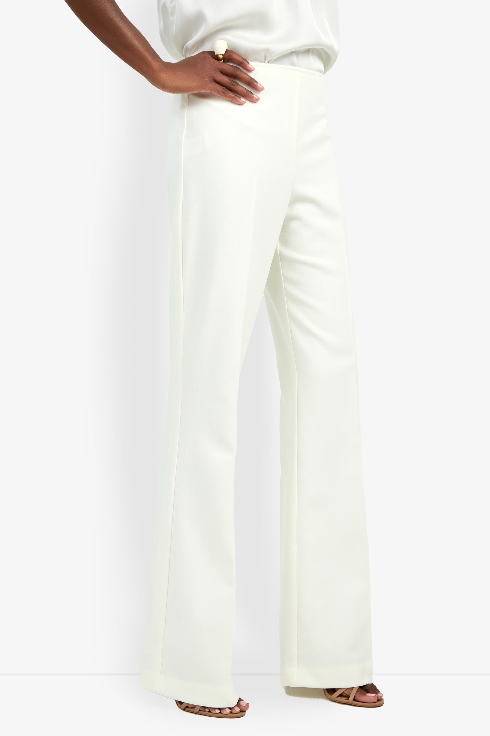 A person wears the CHIMAYO 2 PANT—high-waisted, wide-leg white dress pants made from a polyester-viscose blend—paired with open-toe heels. One hand rests on their hip against a plain white background.