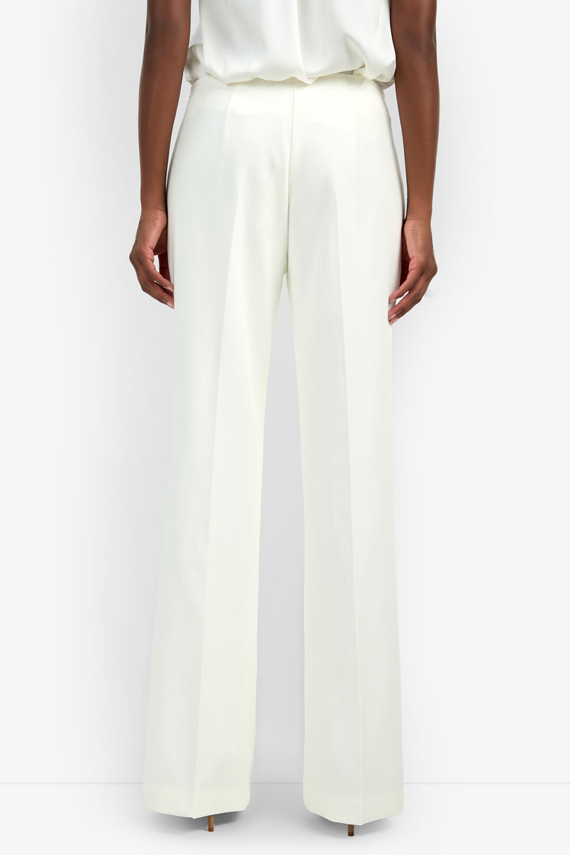 A person stands facing away, wearing the Chimayo Pant—high-waisted, wide-leg white trousers—and a tucked-in white top against a plain white background.