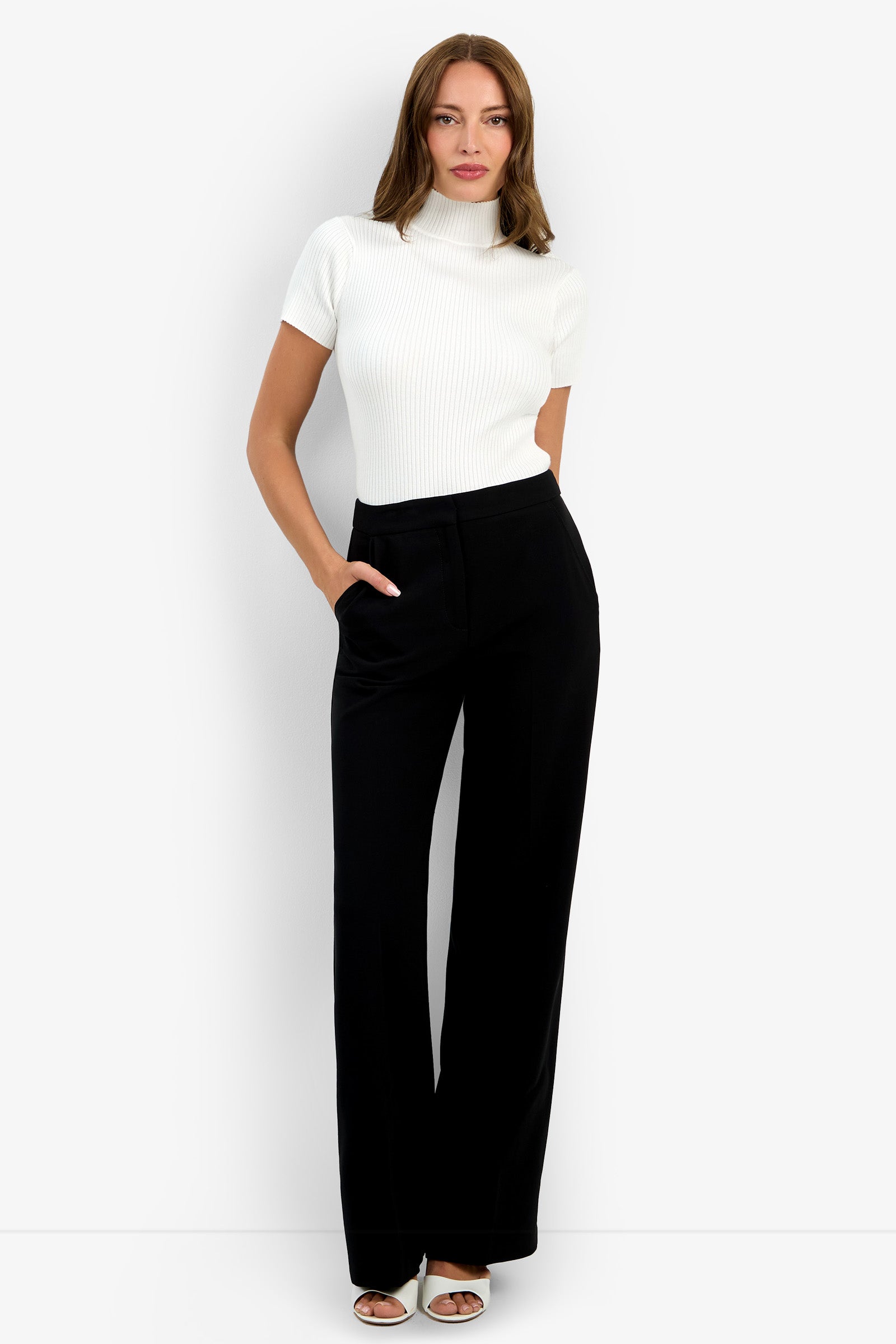 A woman poses against a plain white background, wearing the Sadie Pant—high-waisted black wide-leg pants—with a white short-sleeve ribbed turtleneck top and white open-toe heels, hands in her pockets.