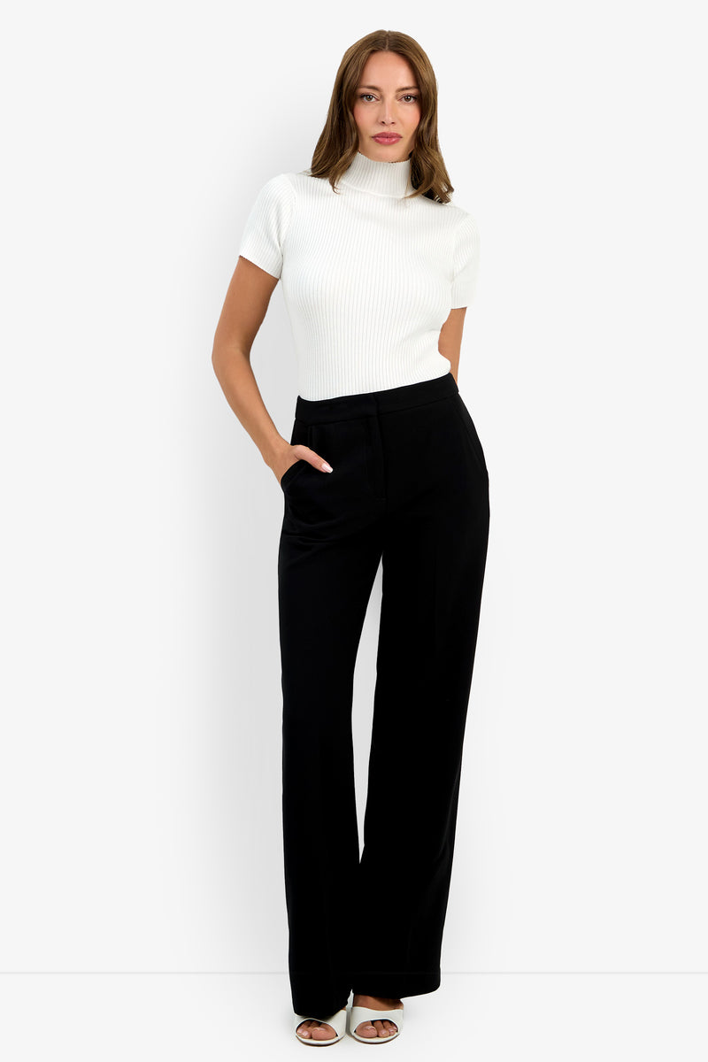 A woman poses confidently against a white background, wearing high-waisted black Sadie Pants, a short-sleeve white turtleneck top, and white open-toe heels. One hand rests in her pocket as she gazes at the camera.