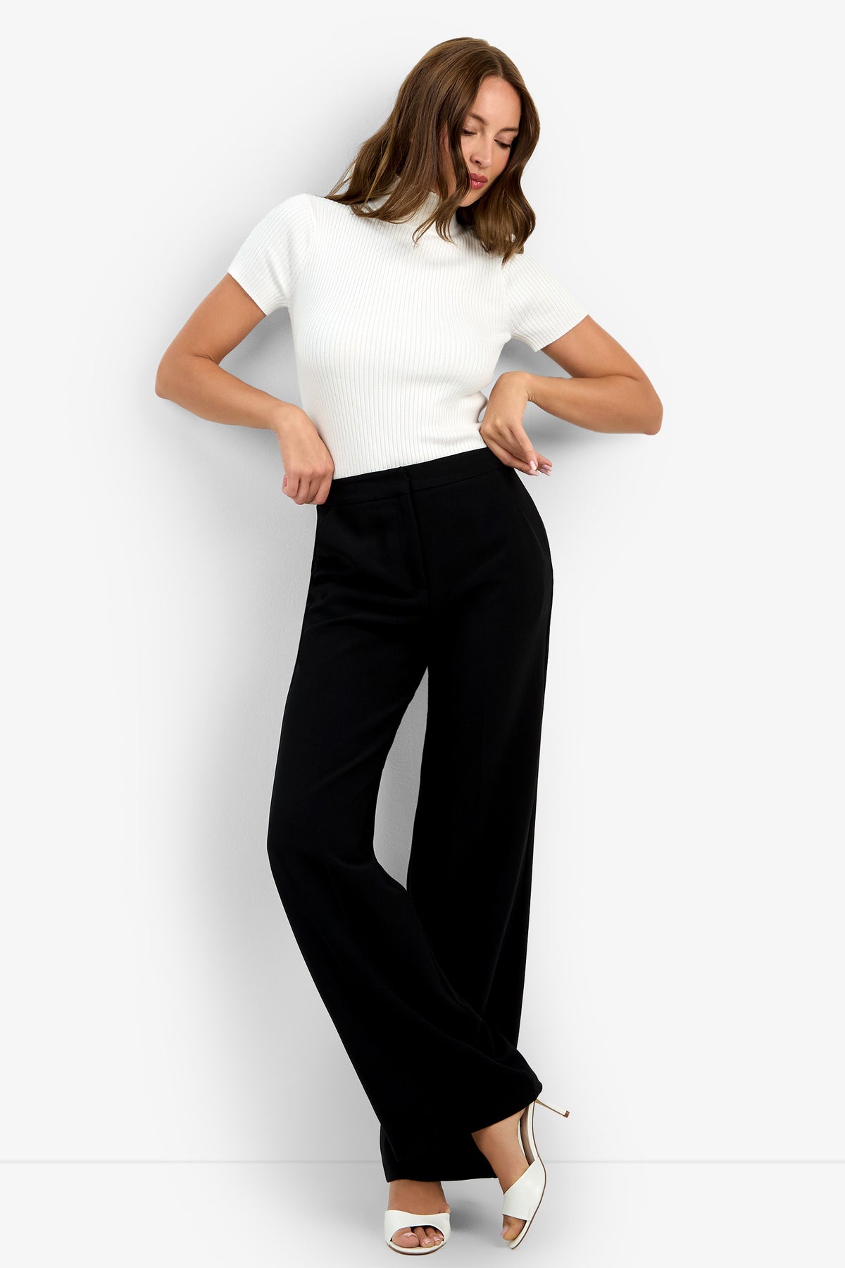 A woman poses against a white backdrop wearing a fitted white short-sleeve top, the high-waisted black Sadie Pant with wide legs, and white heeled sandals. Her arms are slightly bent as she looks downward.