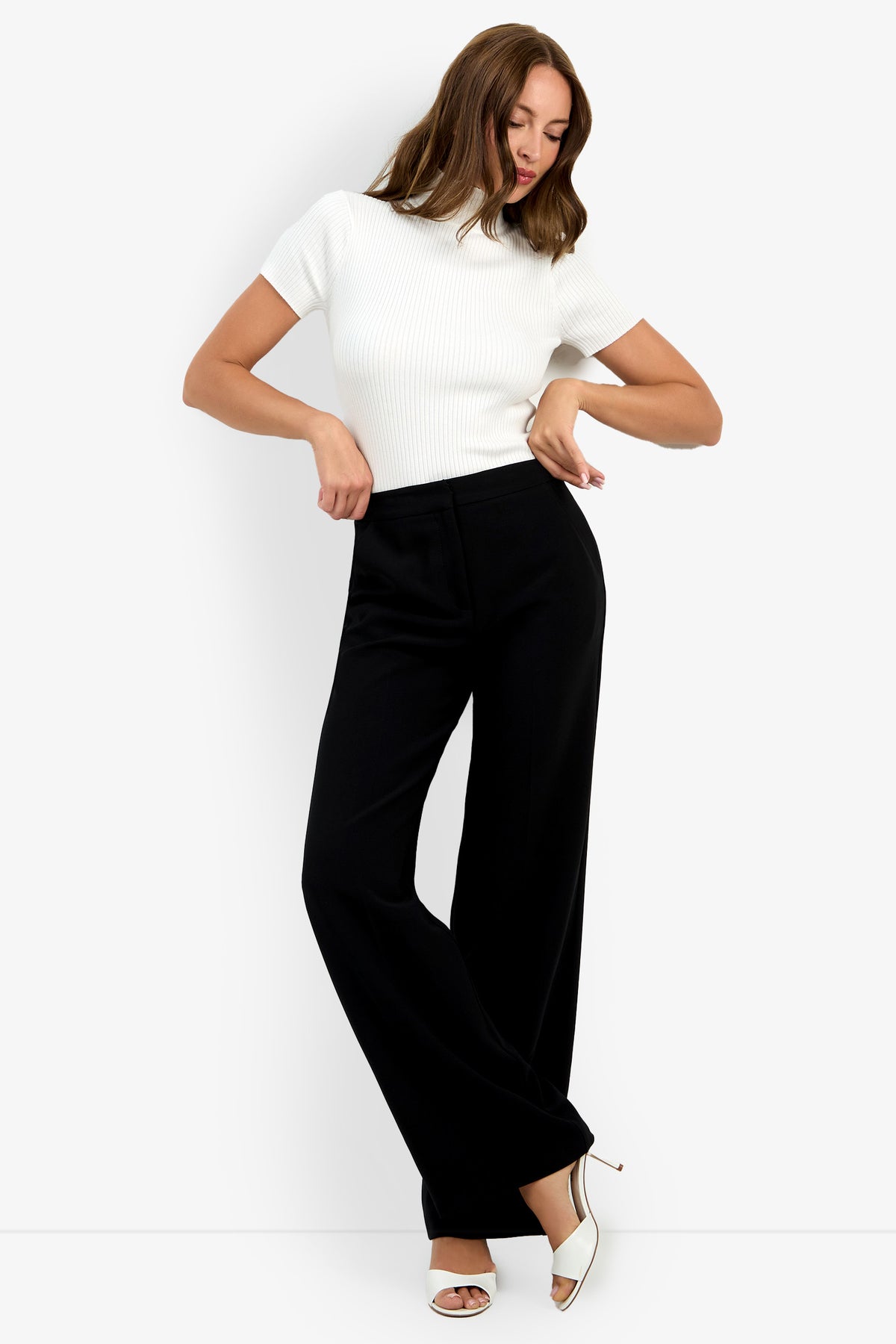 A woman poses against a white background in the Sadie Pant—a pair of black wide-leg pants—styled with a short-sleeved white ribbed top and white open-toe heels. Her hair is loose, arms bent, and her head is slightly tilted.