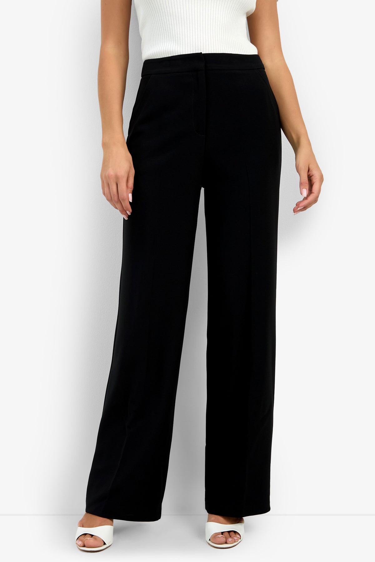 The lower half of a person is shown wearing the Sadie Pant in black, paired with a sleeveless white ribbed top and white open-toe heels, standing against a plain white background.