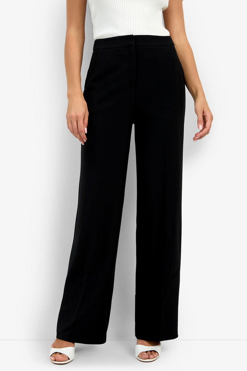 The lower half of a person is shown wearing the Sadie Pant in black, paired with a sleeveless white ribbed top and white open-toe heels, standing against a plain white background.
