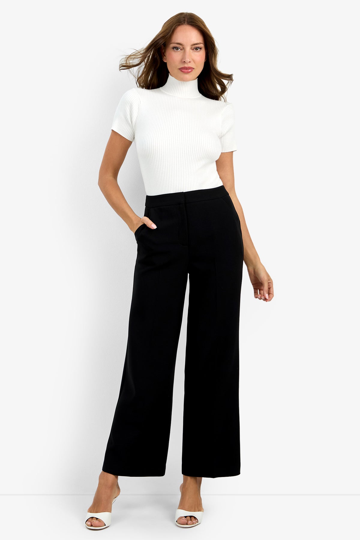 A woman stands against a white background, wearing Sadie Petite Pant—high-waisted, wide-leg black pants—paired with a white short-sleeve turtleneck top and white open-toe heels, one hand in her pocket.