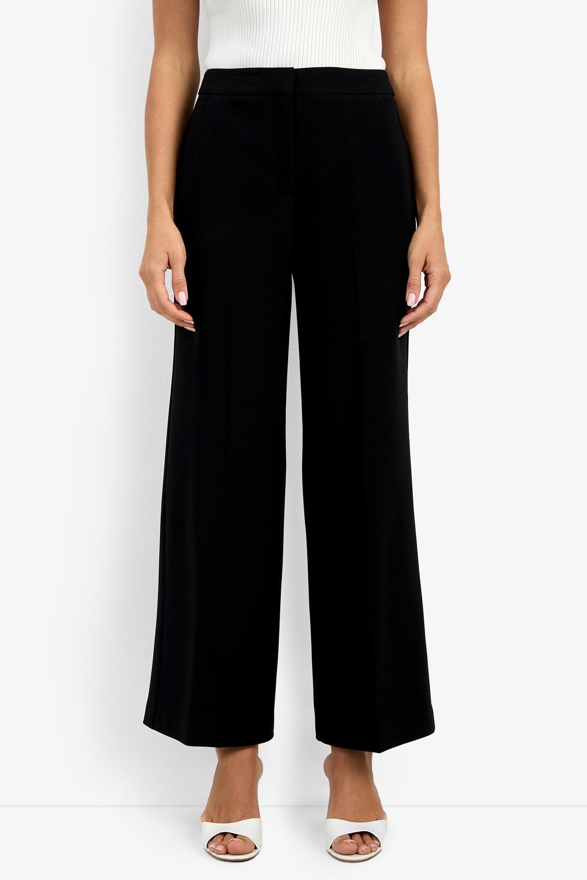 A person wearing the Sadie Petite Pant in black, styled with a white sleeveless top and white open-toe heels, stands against a plain white background.