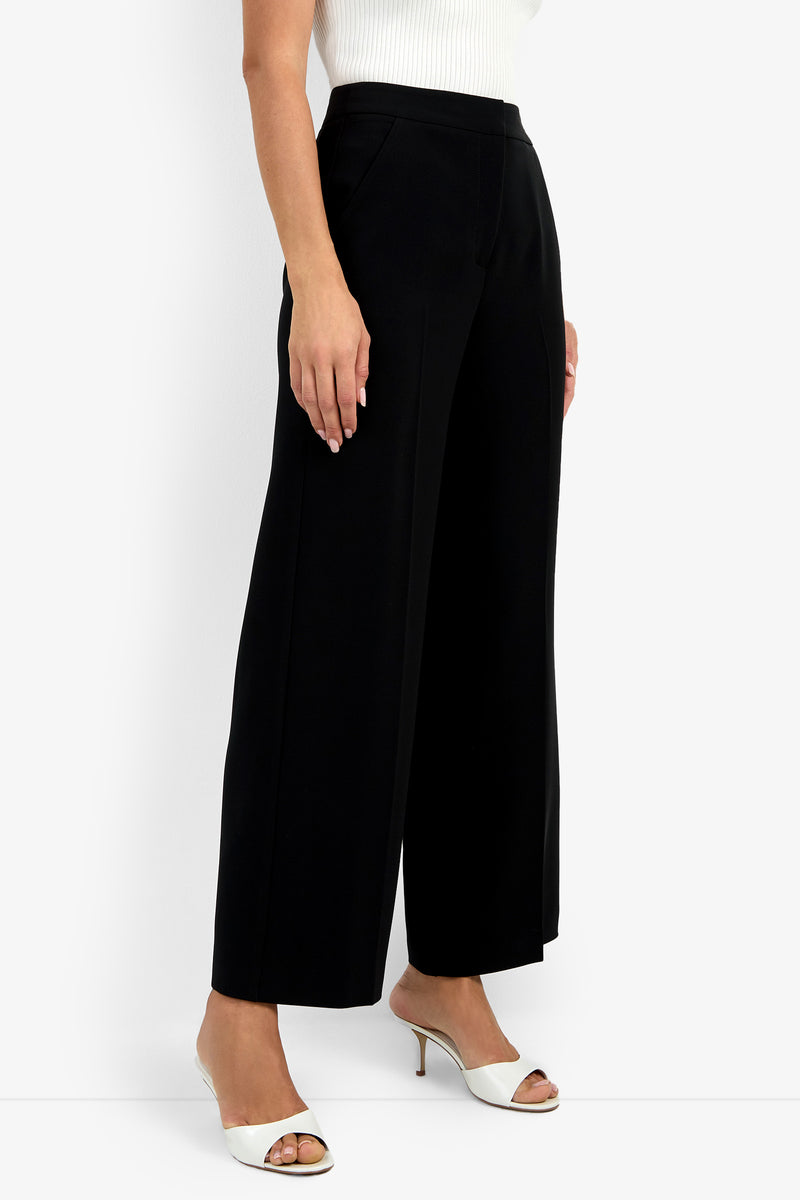 A person wearing Sadie Petite Pant high-waisted black wide-leg trousers, a white ribbed sleeveless top, and white open-toe heeled sandals stands against a plain white background.