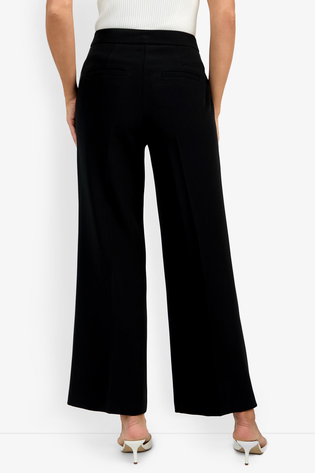 A person wearing the Sadie Petite Pant in black, styled with white heeled sandals and a sleeveless white top. Shown from the back, the pants feature two welt pockets.