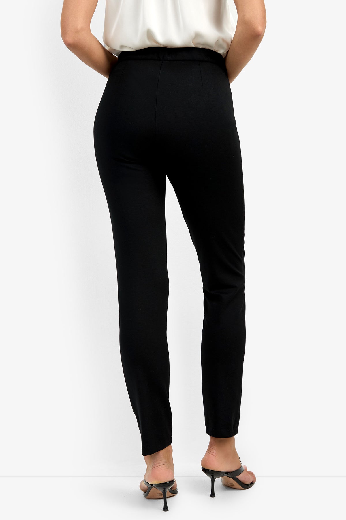 Someone wearing the Honey Pant in black, a white sleeveless top, and black heeled sandals stands with their back to the camera against a plain white background.
