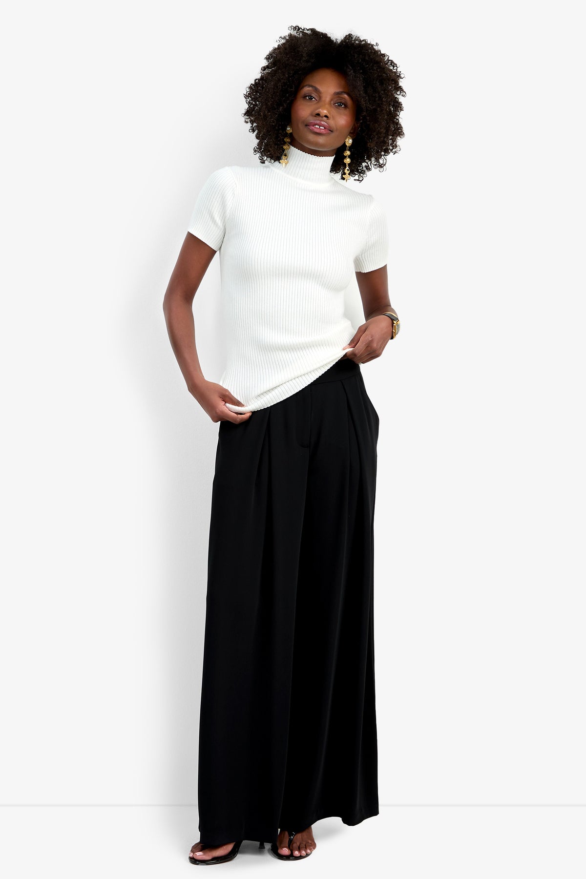 A woman with curly hair and gold earrings stands against a white background, slightly smiling while wearing the Saige Pant—wide-leg black pants—paired with a short-sleeve white turtleneck sweater and black sandals.