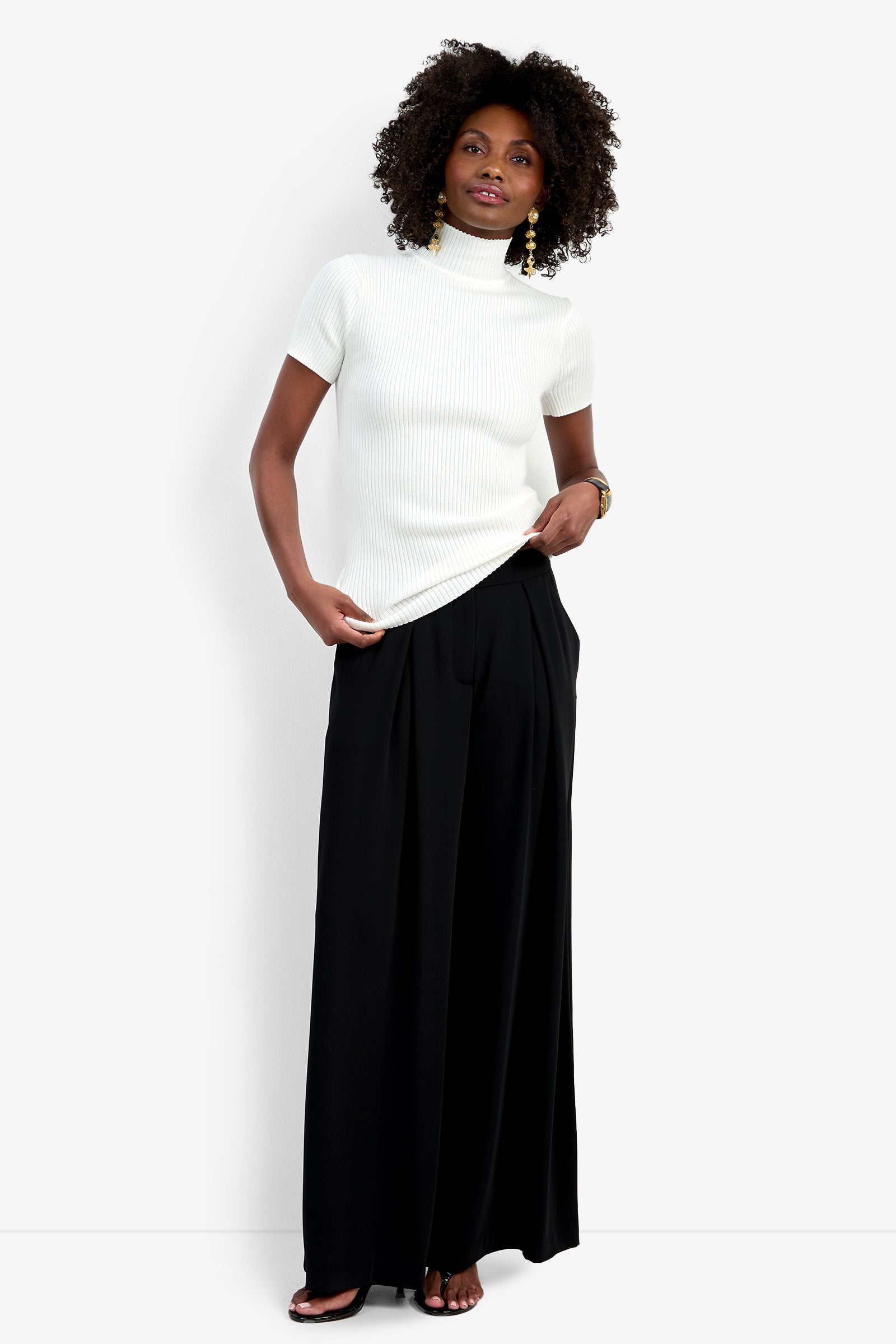 A woman with curly hair and gold earrings stands against a white background, slightly smiling while wearing the Saige Pant—wide-leg black pants—paired with a short-sleeve white turtleneck sweater and black sandals.