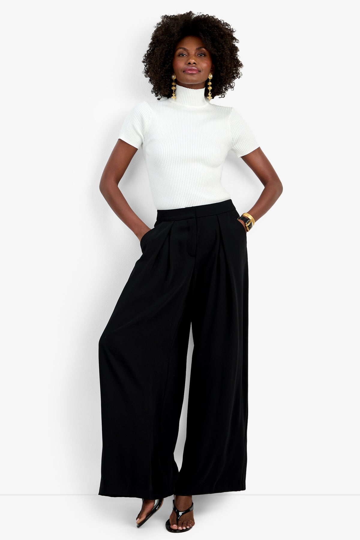 A woman stands confidently with hands in her pockets, wearing the Saige Pant—wide-leg, high-waisted black pants—paired with a white short-sleeve turtleneck sweater, heeled sandals, gold earrings, and a bracelet against a plain background.