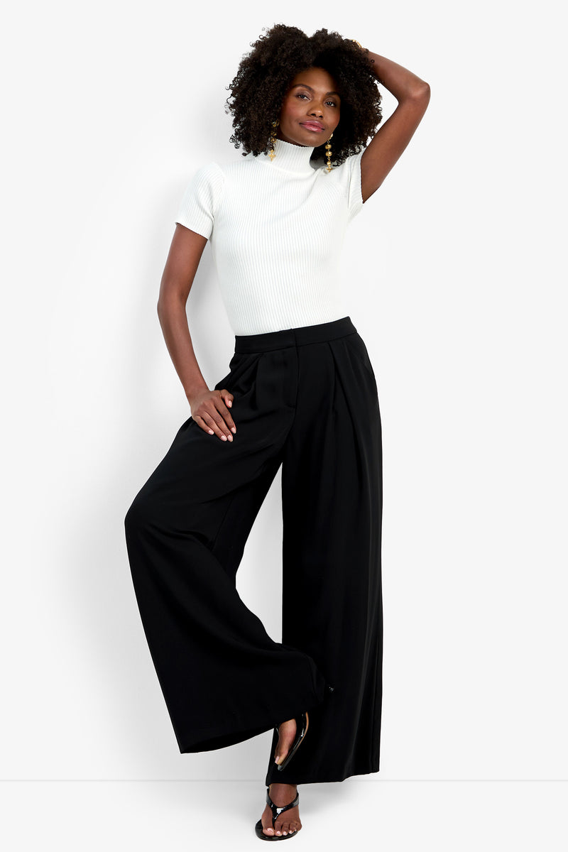 A woman poses confidently in the Saige Pant—wide-leg and black—styled with a white short-sleeve turtleneck top and black heeled sandals, her hand resting on her head against a plain white backdrop.