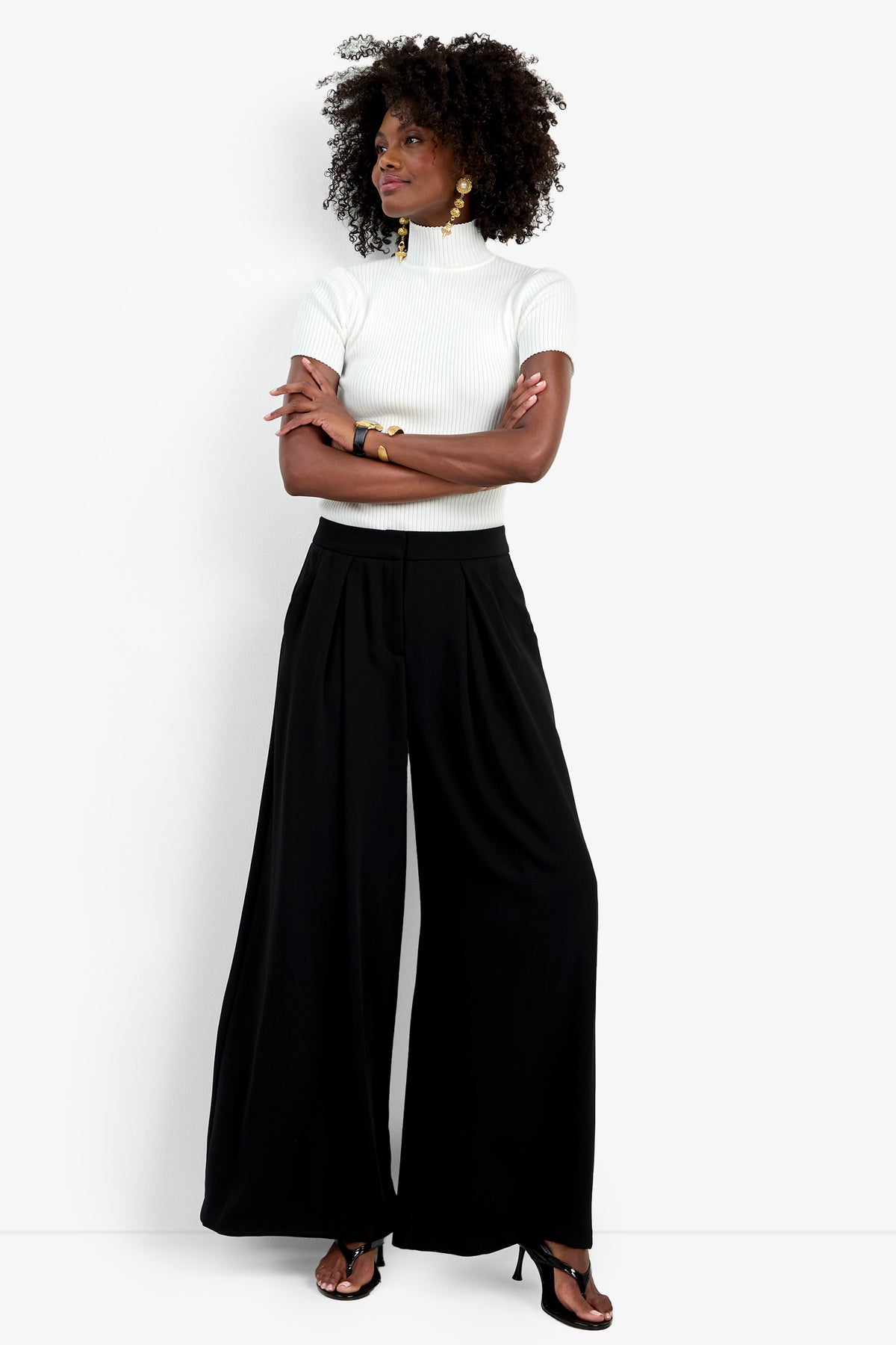 A woman with curly hair stands with arms crossed, wearing wide-leg black Saige Pants, a white short-sleeve turtleneck top, black heeled sandals, and gold earrings. She looks to the side against a plain white background.